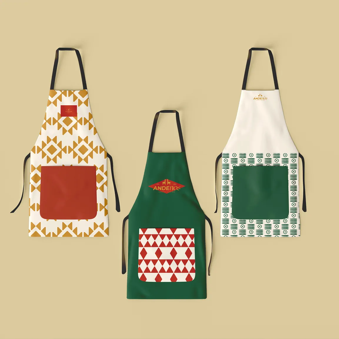 Trois tabliers à motifs, l’un ocre et blanc, l’un vert avec le logo d’Andenos, l’autre blanc cassé et vert, posés sur un fond sable.