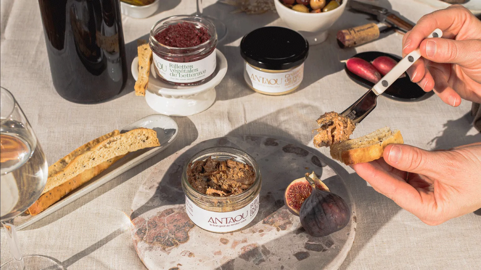 Une main tend un biscuit vers un pot de rillettes Antaou, au milieu d’une table : figues, olives, verres de vin et planches.
