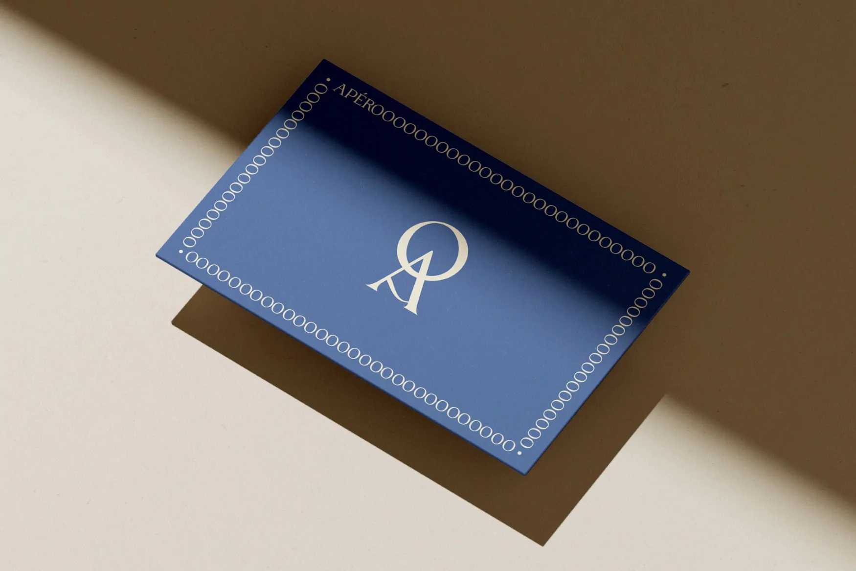 Une carte bleu marine ornée d’une frise blanche et du monogramme, posée sur un fond ocre.