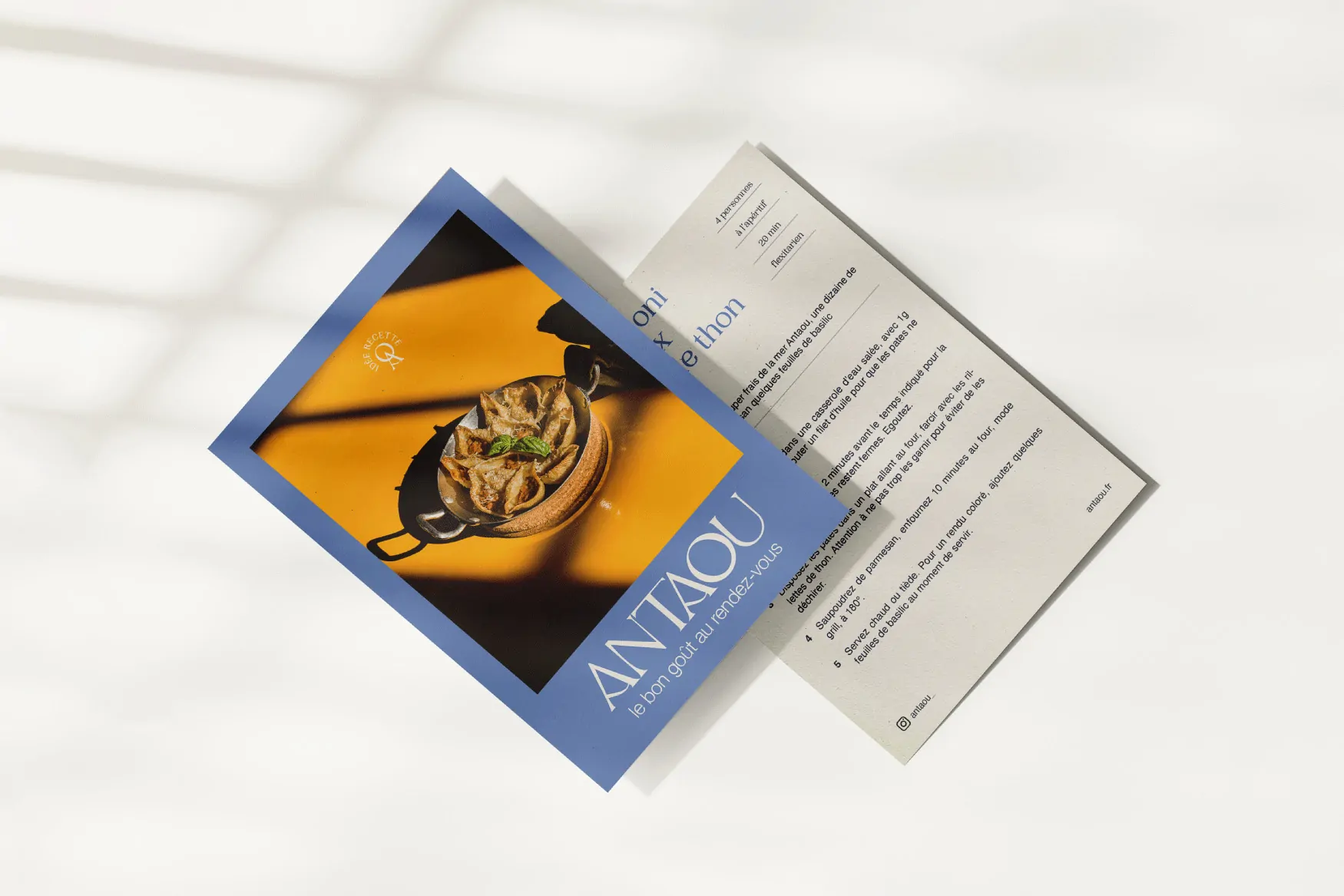 Une carte postale de recette bleue posée sur un fond blanc, à côté d’un plat sur fond jaune.