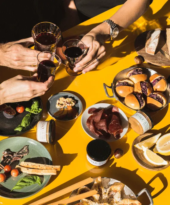 Des mains trinquent avec des verres de vin au-dessus d’une table jaune chargée de tapas.