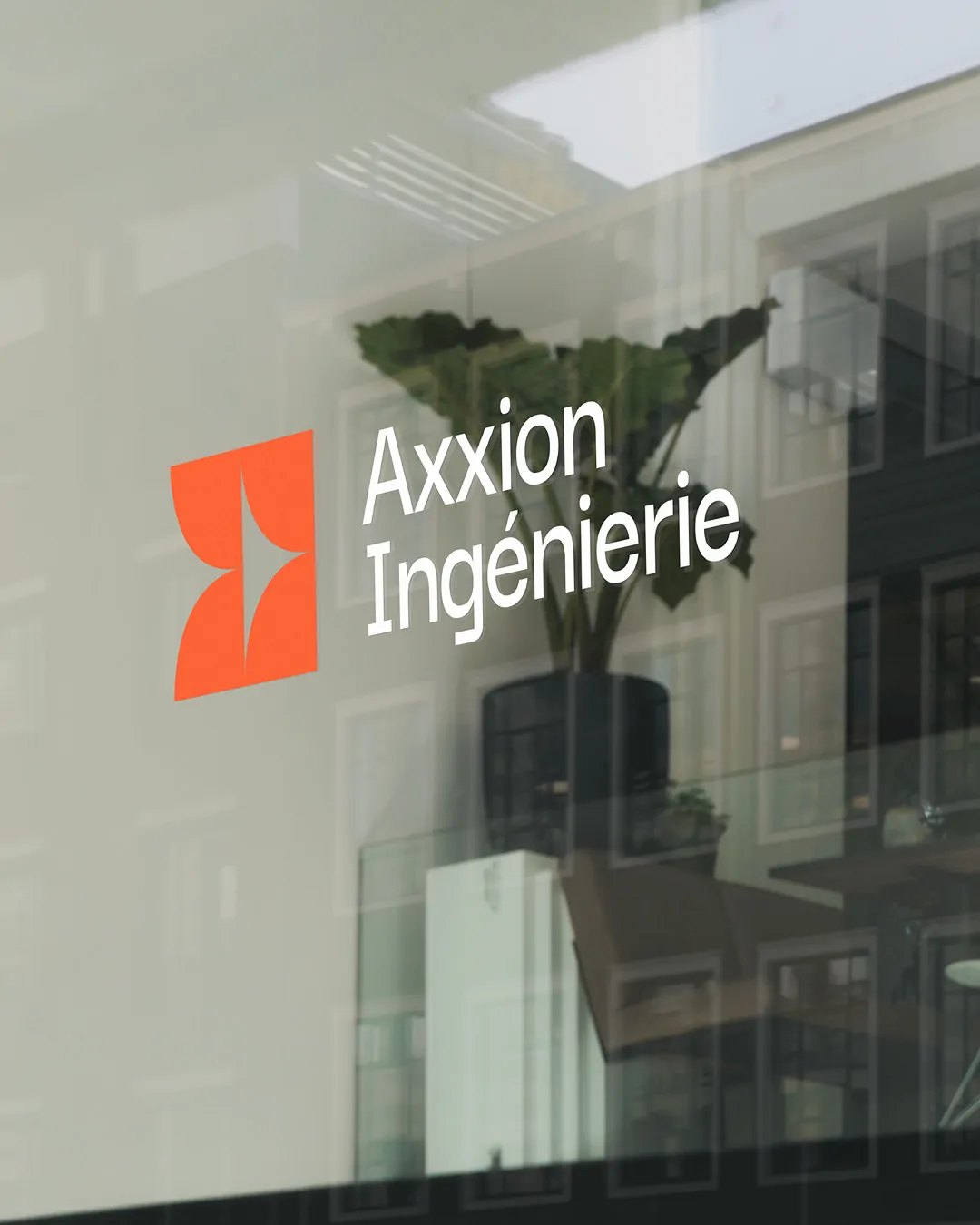 Le logo Axxion Ingénierie sur une vitre qui reflète une façade et une plante.