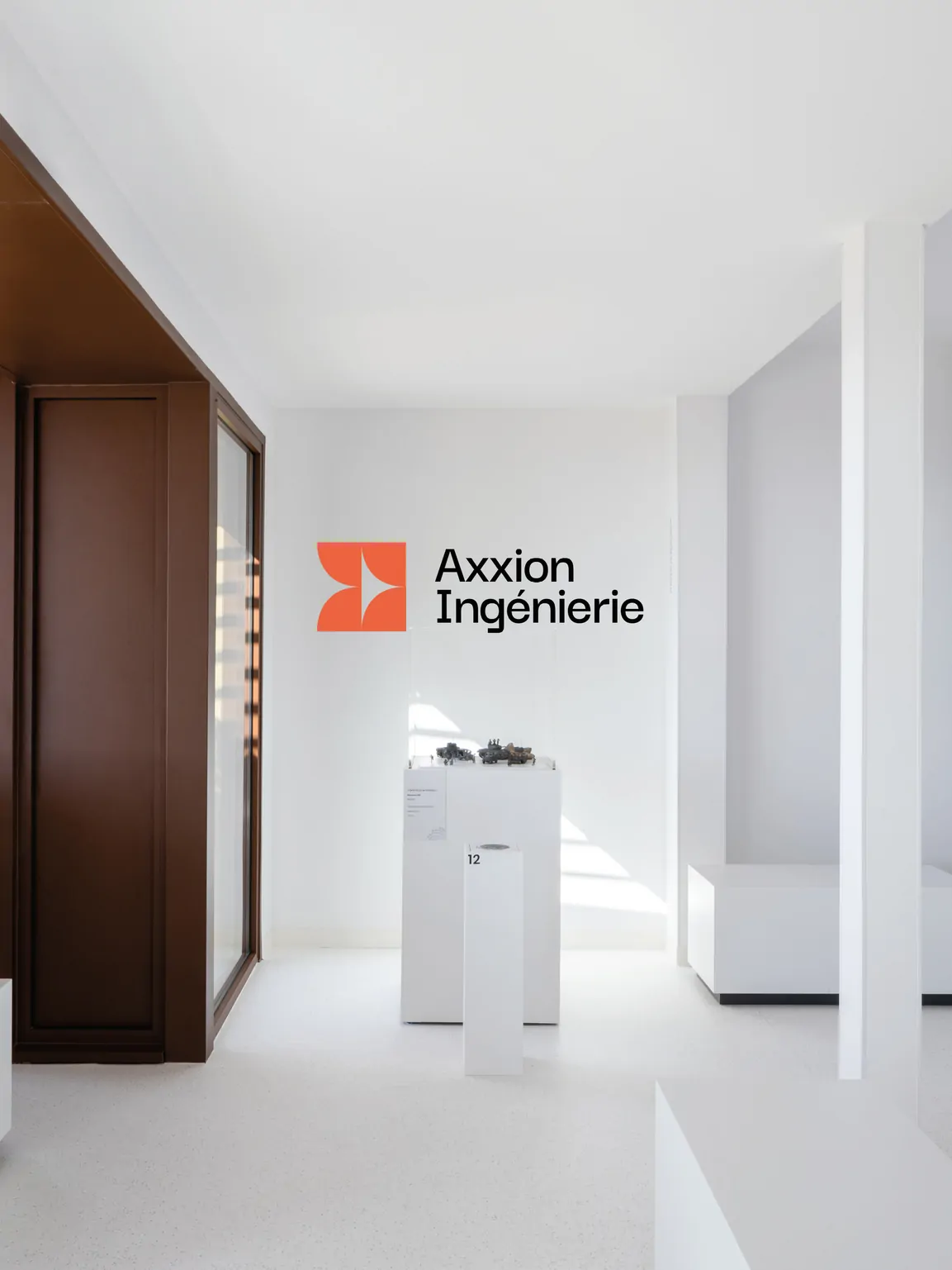Une salle d’exposition blanche, un mur portant le logo Axxion Ingénierie, une paroi brune et un piédestal avec une maquette.