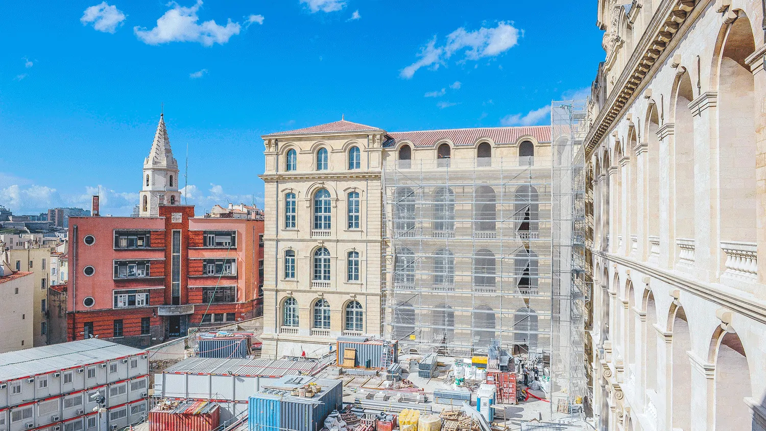 Un grand bâtiment de pierre sous échafaudage au cœur de Marseille, entre des immeubles et un clocher.