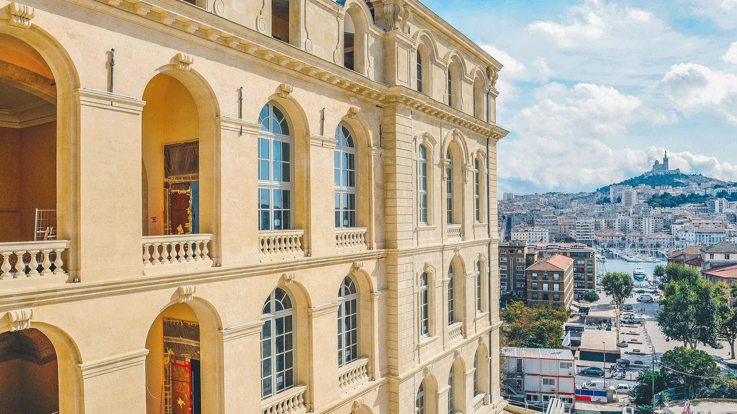 Les arcades d’un monument de pierre dorée, avec la ville de Marseille et la colline de Notre-Dame de la Garde à l’horizon.