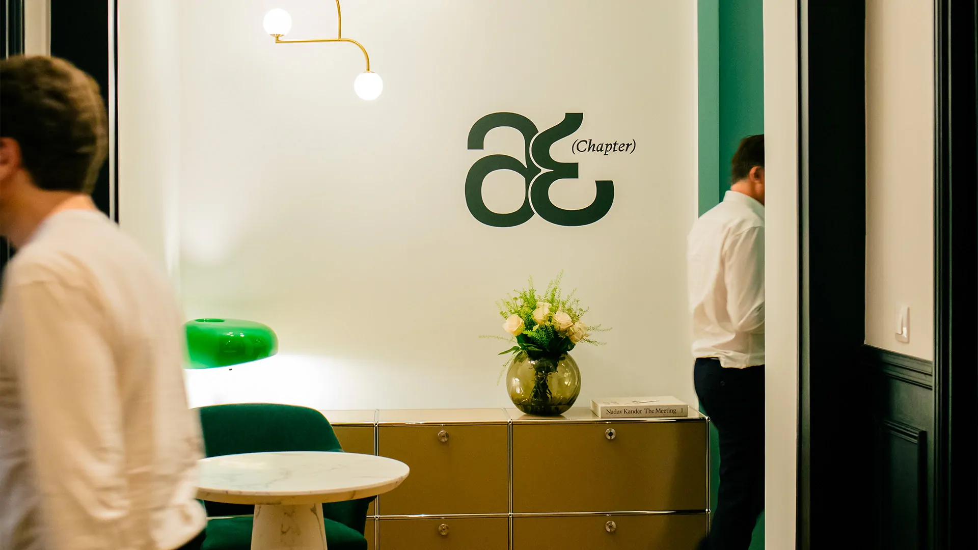 Au bureau du cabinet, le monogramme « æ (Chapter) » en vert sur un mur blanc, un bouquet de roses, un buffet en laiton et deux silhouettes en chemise blanche.
