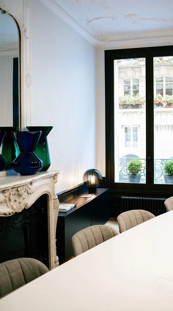 Un salon du cabinet : une cheminée en marbre, des vases verts, une lampe-globe et une grande table blanche devant deux fenêtres.