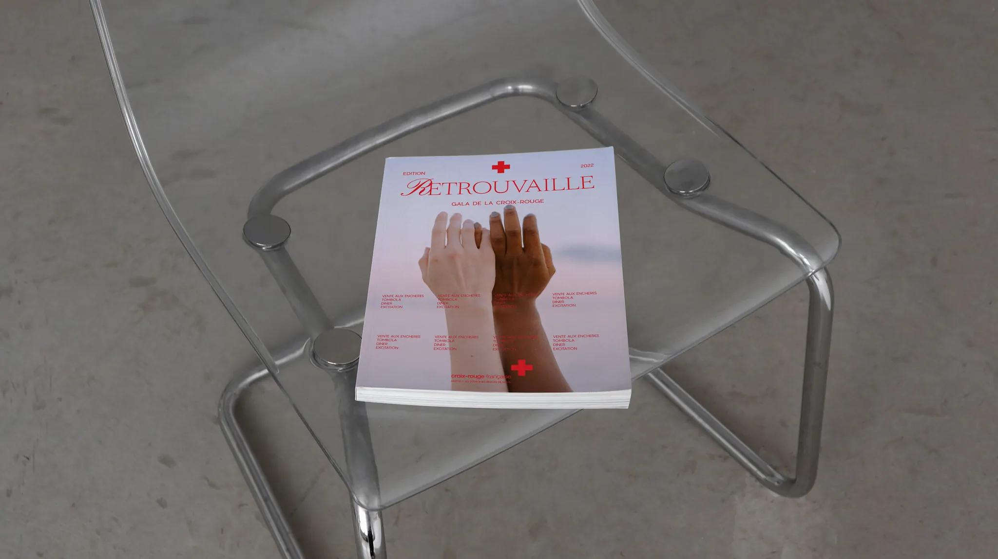 Le catalogue de l’événement, fermé, posé sur une chaise transparente : sur la couverture, deux mains, une claire et une foncée.