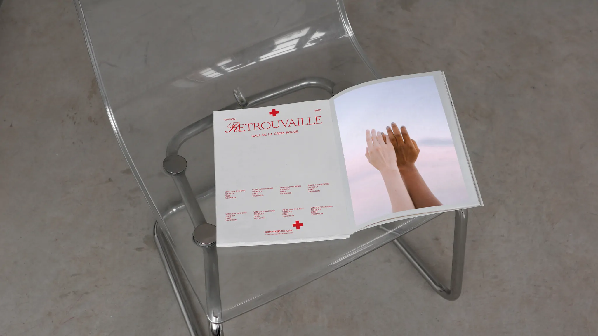 Le catalogue ouvert sur la chaise transparente, ses pages claires et la photo des deux mains.