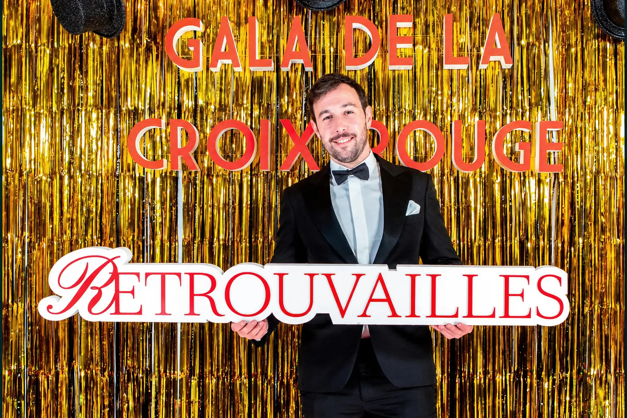 Un invité en smoking et nœud papillon sourit en tenant l’enseigne « Retrouvailles », devant un rideau doré et les lettres « Gala de la Croix-Rouge ».