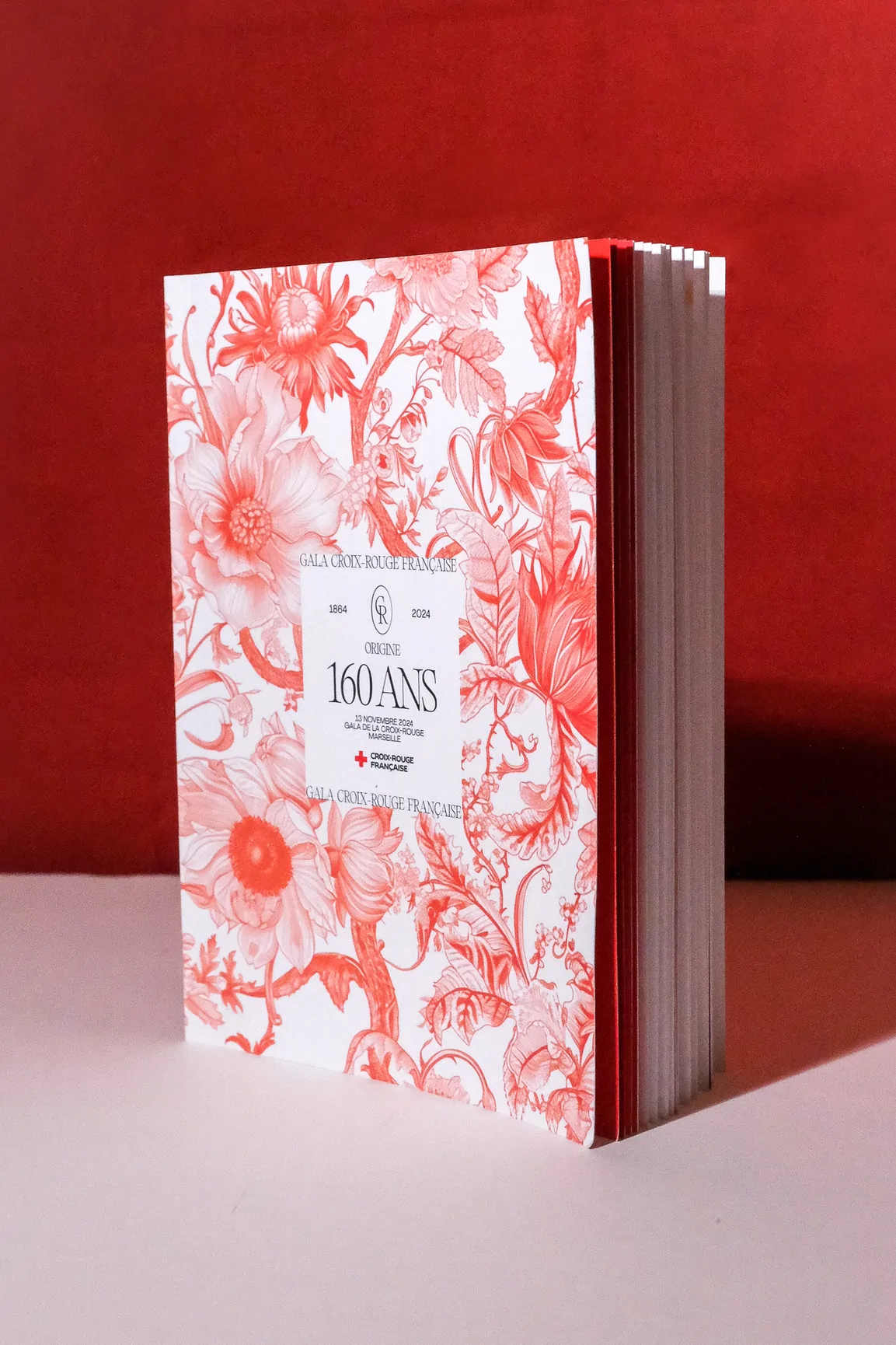 Le catalogue du gala, debout contre un mur rouge : sa couverture porte le motif floral rouge et blanc et un cartouche « Origine 160 ans ».
