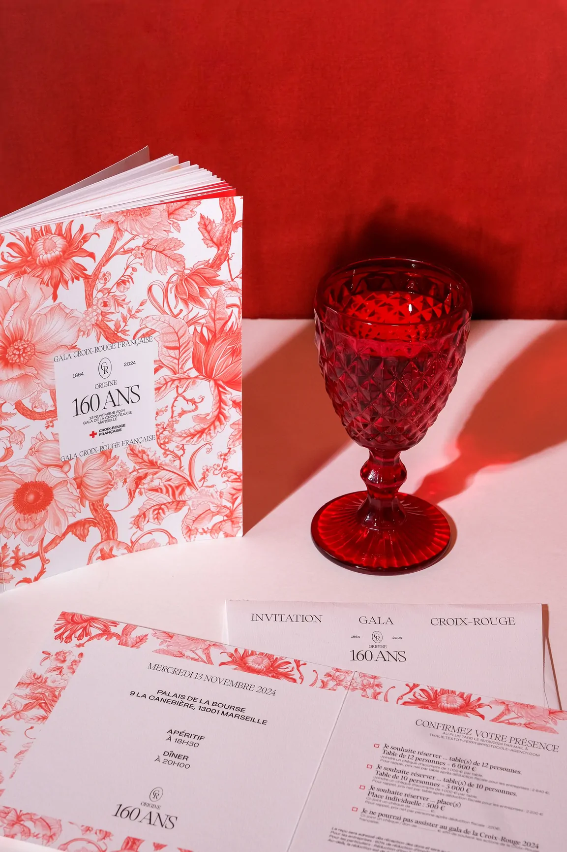 Le catalogue posé près d’un verre en cristal rouge, devant le programme de la soirée, ouvert et bordé du motif floral.