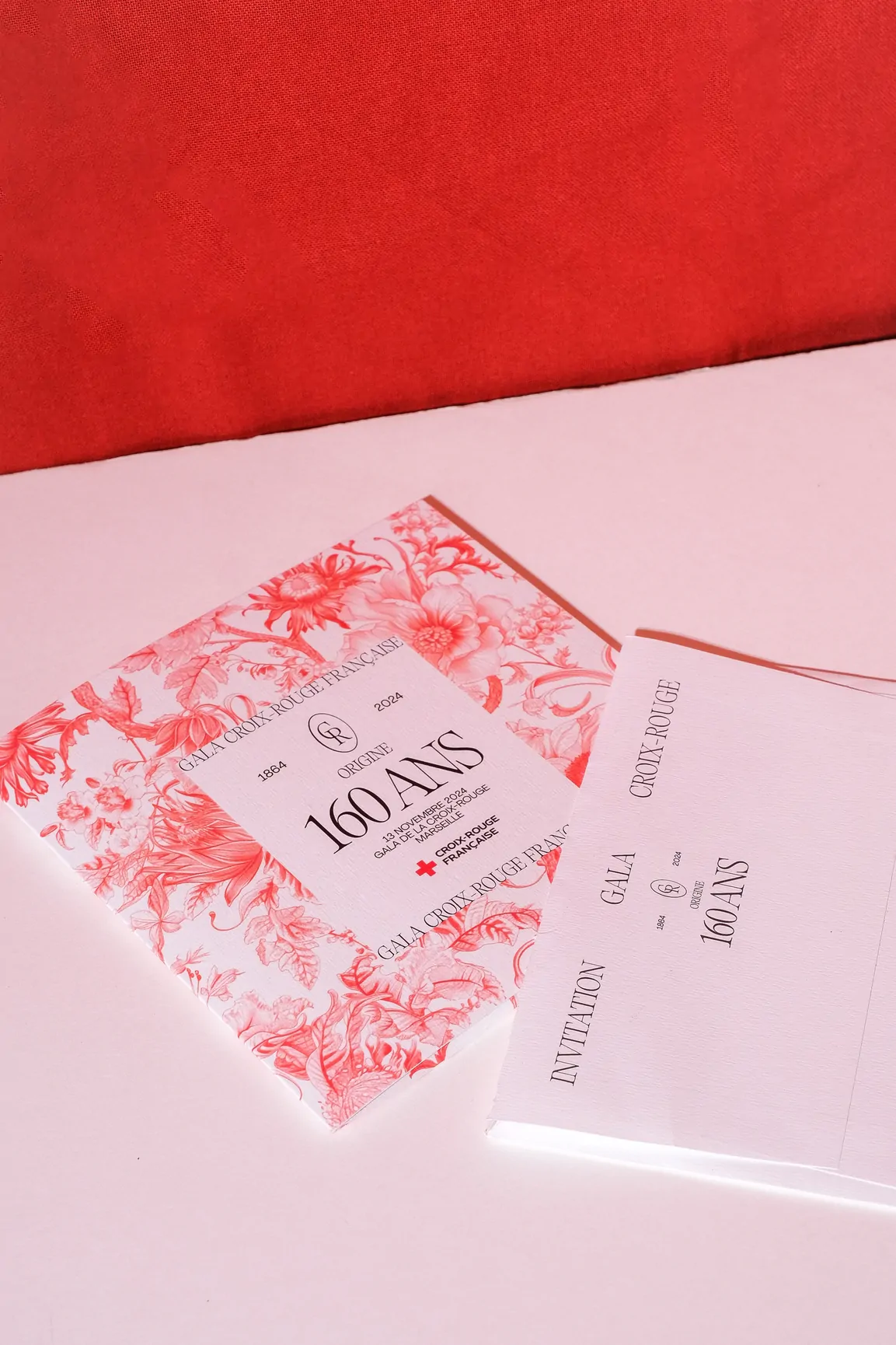 L’invitation au gala, couverte du motif floral, posée sur son enveloppe blanche, sur un fond rouge et rose.