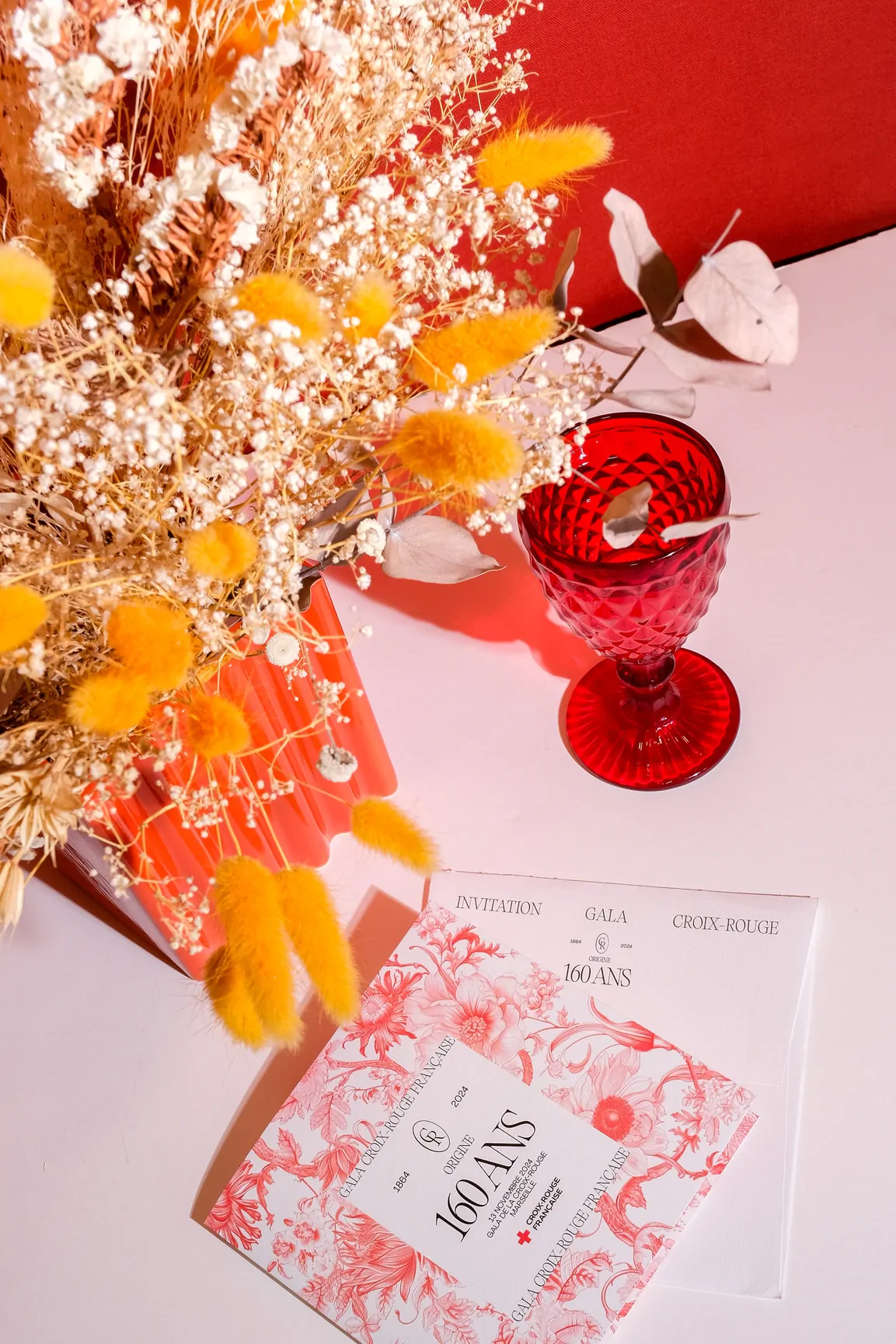L’invitation posée à côté d’un bouquet de fleurs séchées jaunes et orangées et d’un verre rouge.