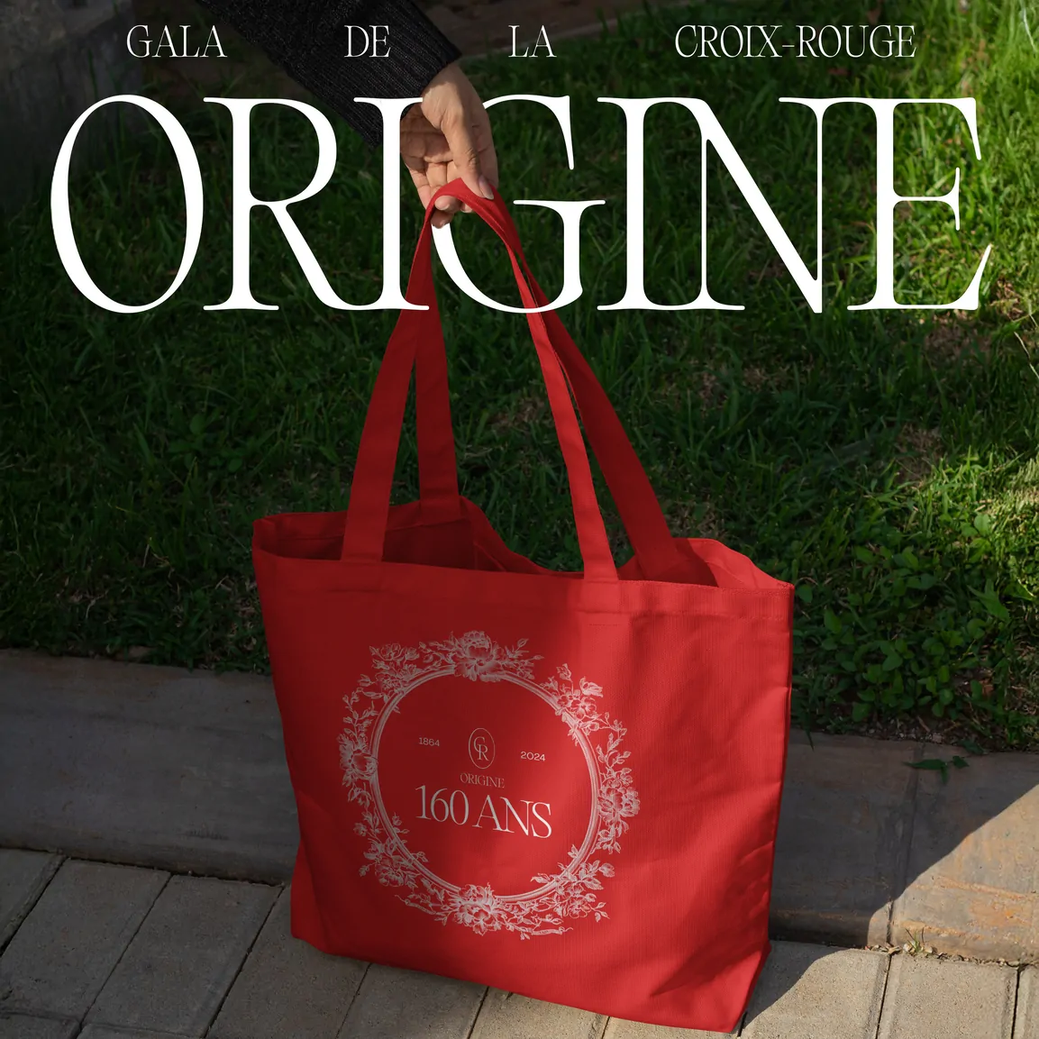 Un tote bag rouge portant l’emblème « Origine 160 ans » en blanc, suspendu devant une pelouse, sous le mot « Origine » en grandes lettres blanches.