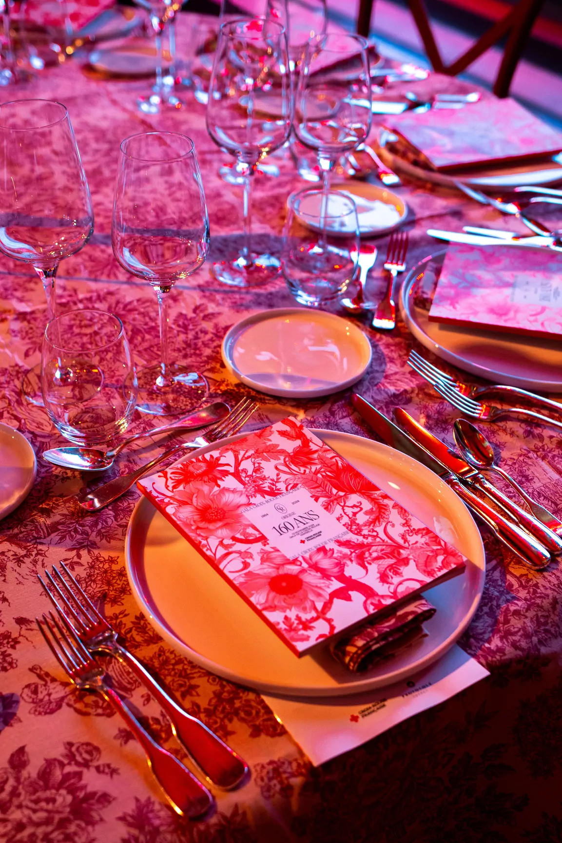 Une table dressée, assiettes et verres sur une nappe imprimée, avec le menu aux couleurs du gala, en lumière rose.