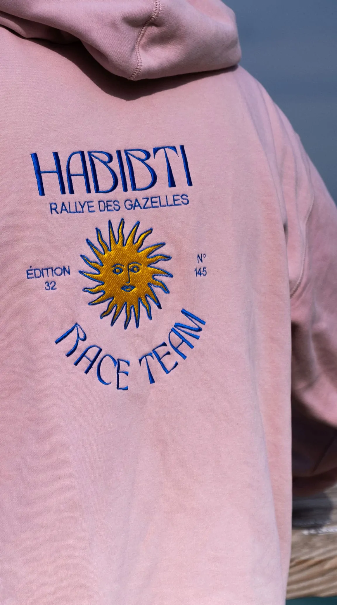 Un sweat rose dont le dos porte le sceau Habibti Race Team et un soleil, devant la mer.