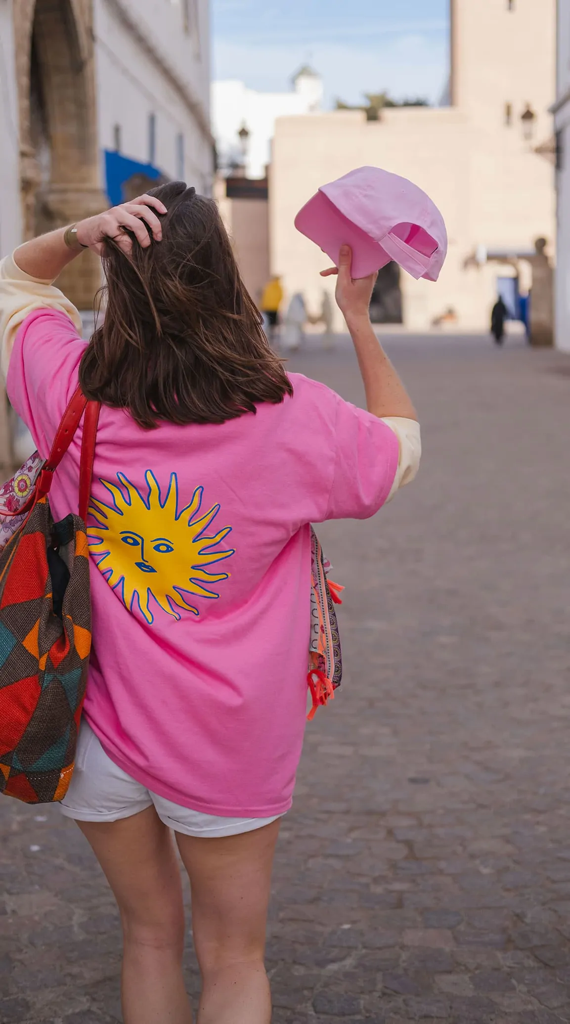 Une coéquipière de dos, en tee-shirt rose au soleil jaune, dans une ruelle d’Essaouira, une casquette rose à la main.