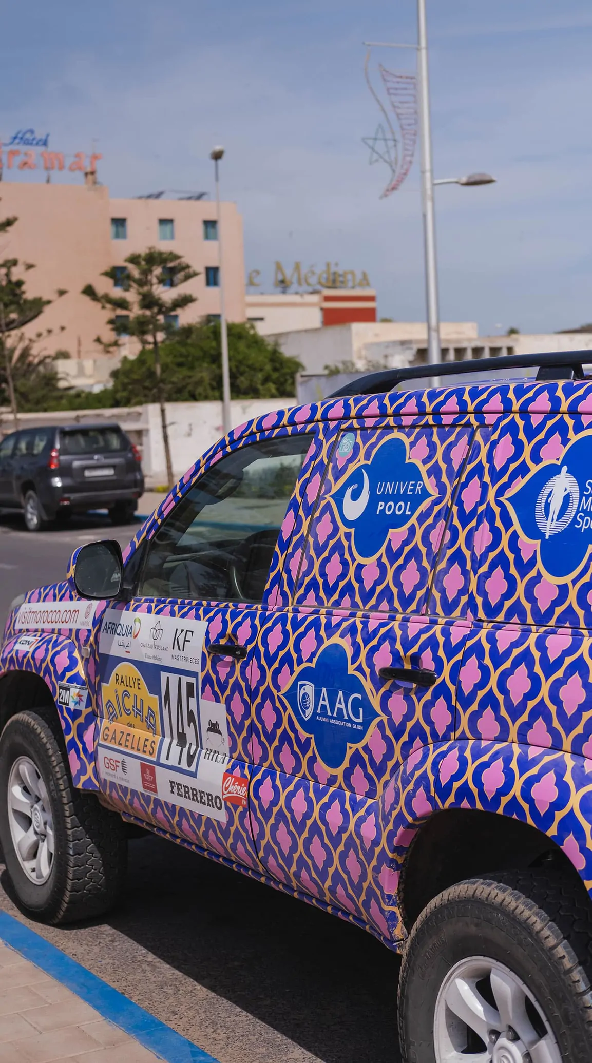 Le 4x4 de l’équipage 145, entièrement recouvert d’un motif géométrique violet et rose, garé devant des immeubles blancs.