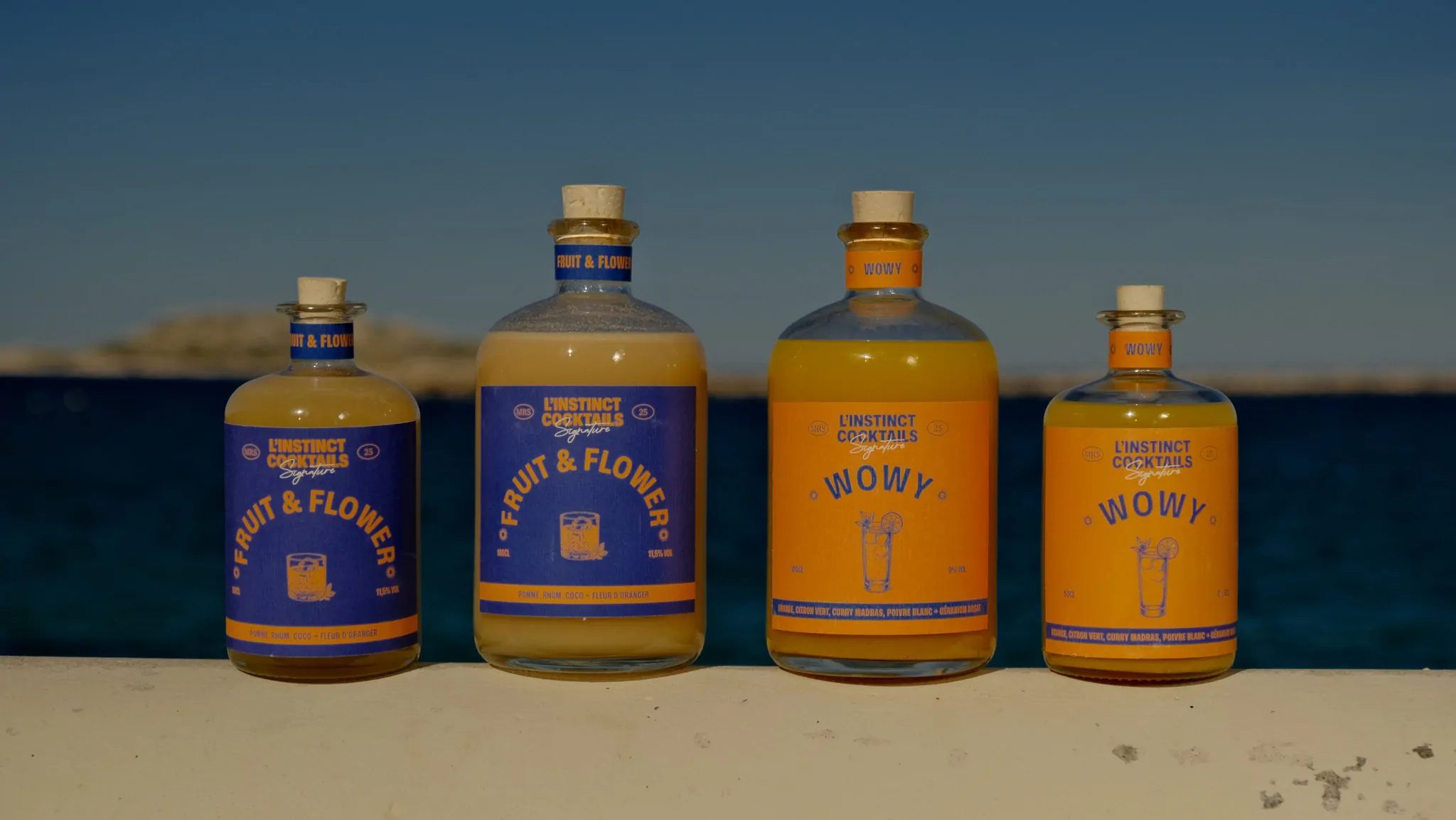 Quatre bouteilles de la gamme alignées sur le sable, la mer et le ciel du soir derrière.