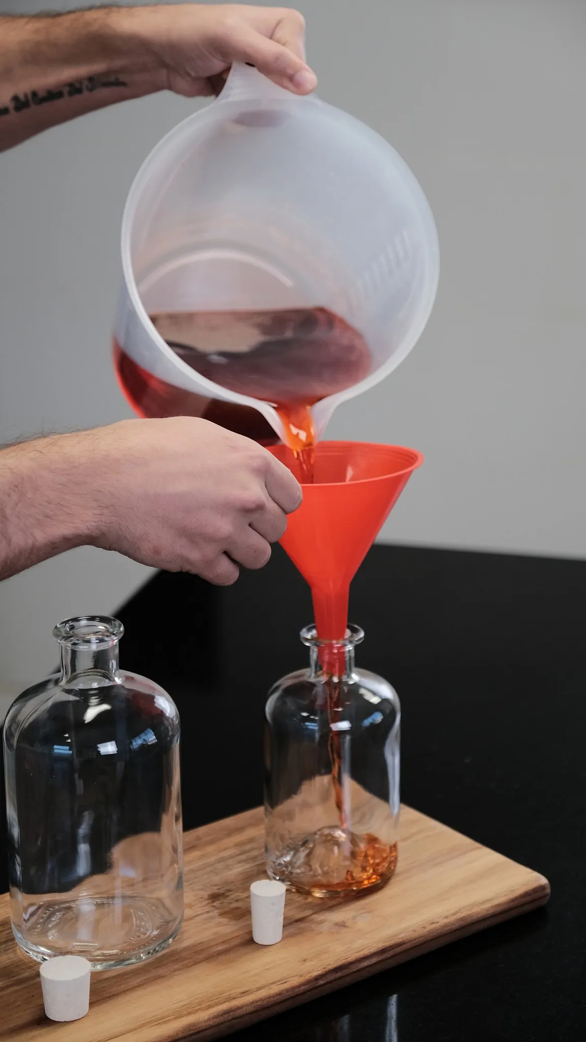 Des mains versent un cocktail rouge d’un broc dans une bouteille à l’aide d’un entonnoir orange, sur une planche en bois.