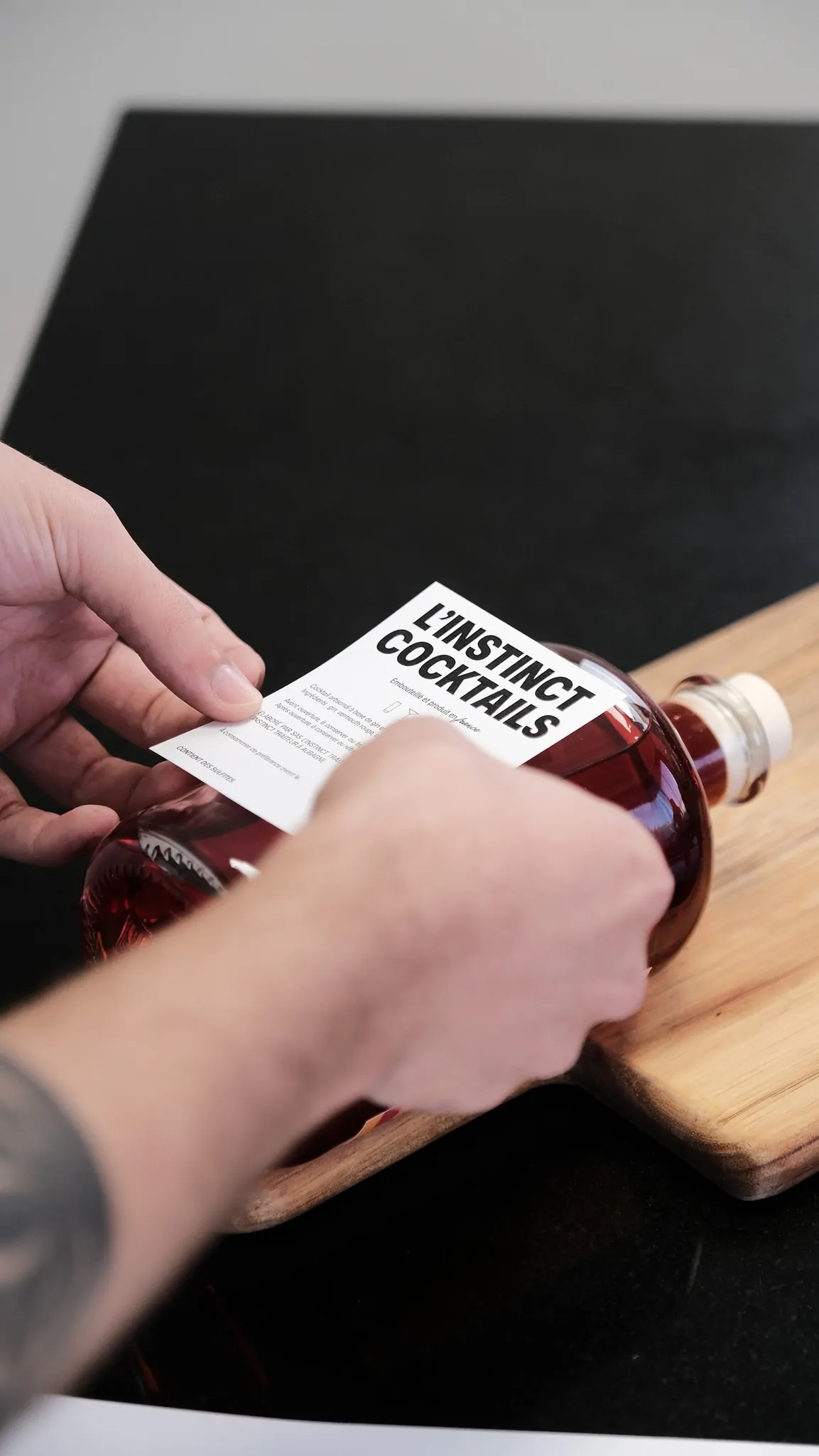 Des mains collent l’étiquette sur une bouteille posée sur une planche en bois.