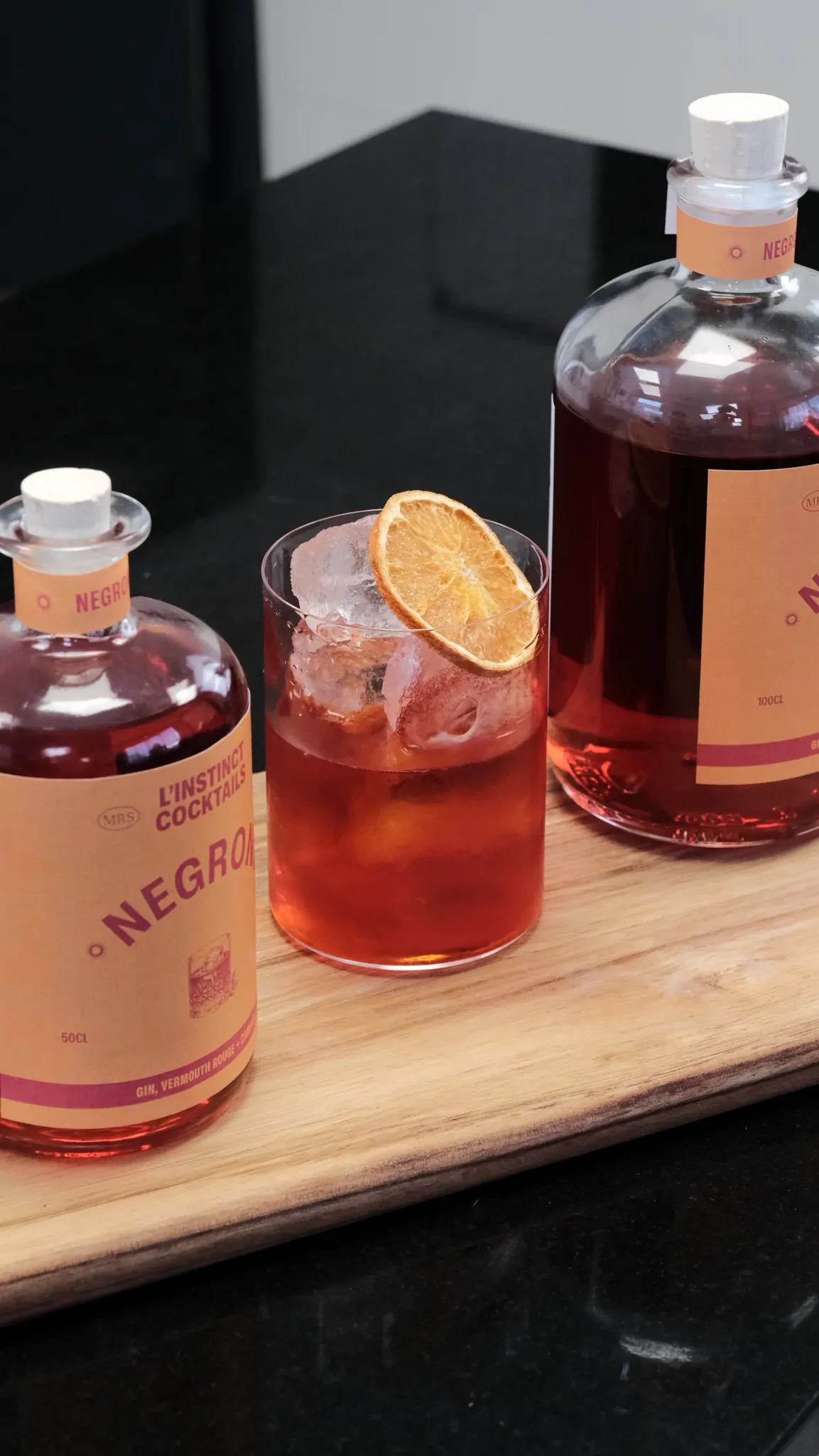 Deux bouteilles de Négroni et un verre garni d’une tranche d’orange sur une planche en bois.