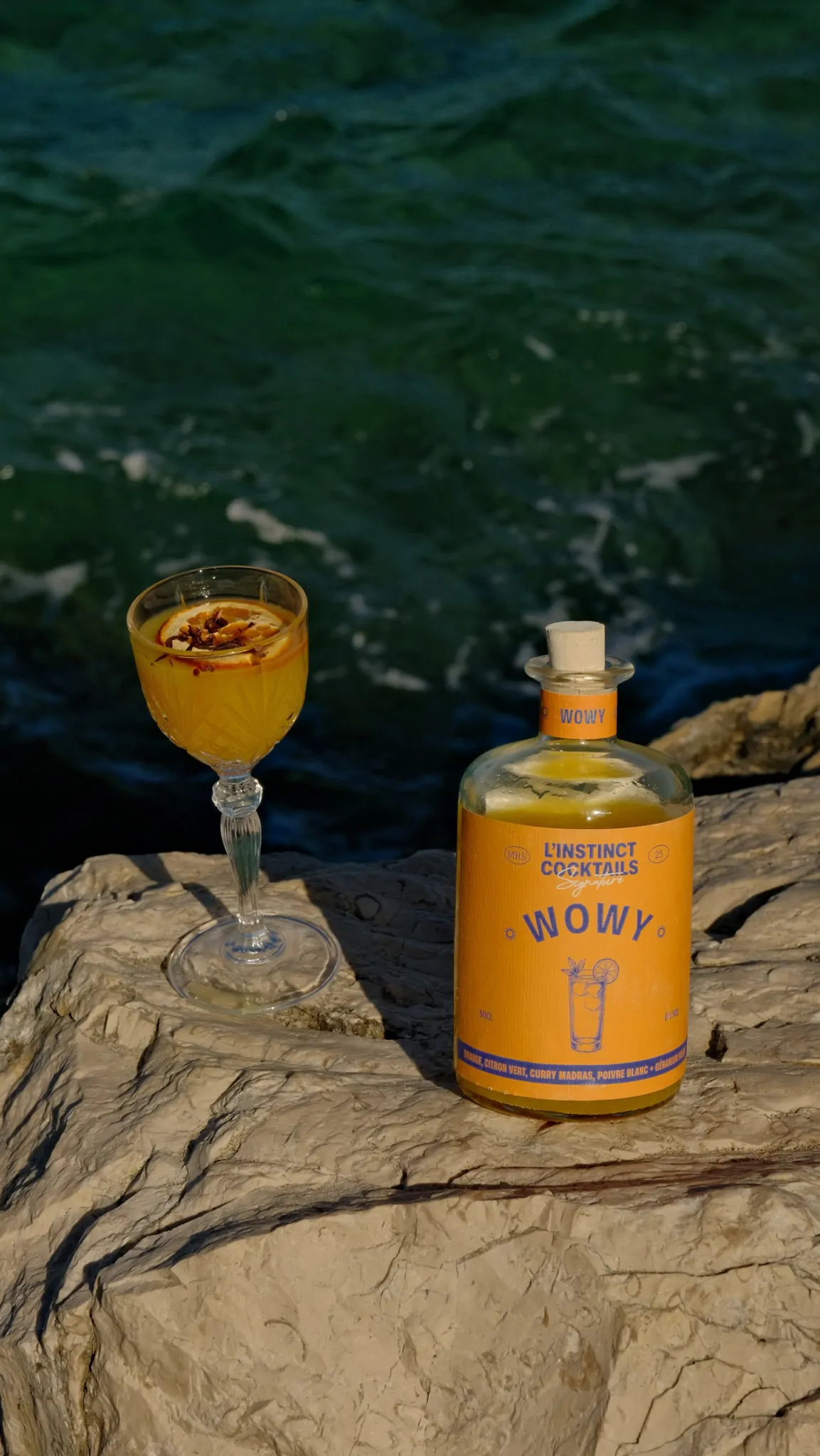 La bouteille Wowy et un verre de cocktail posés sur un rocher, au-dessus d’une eau turquoise.