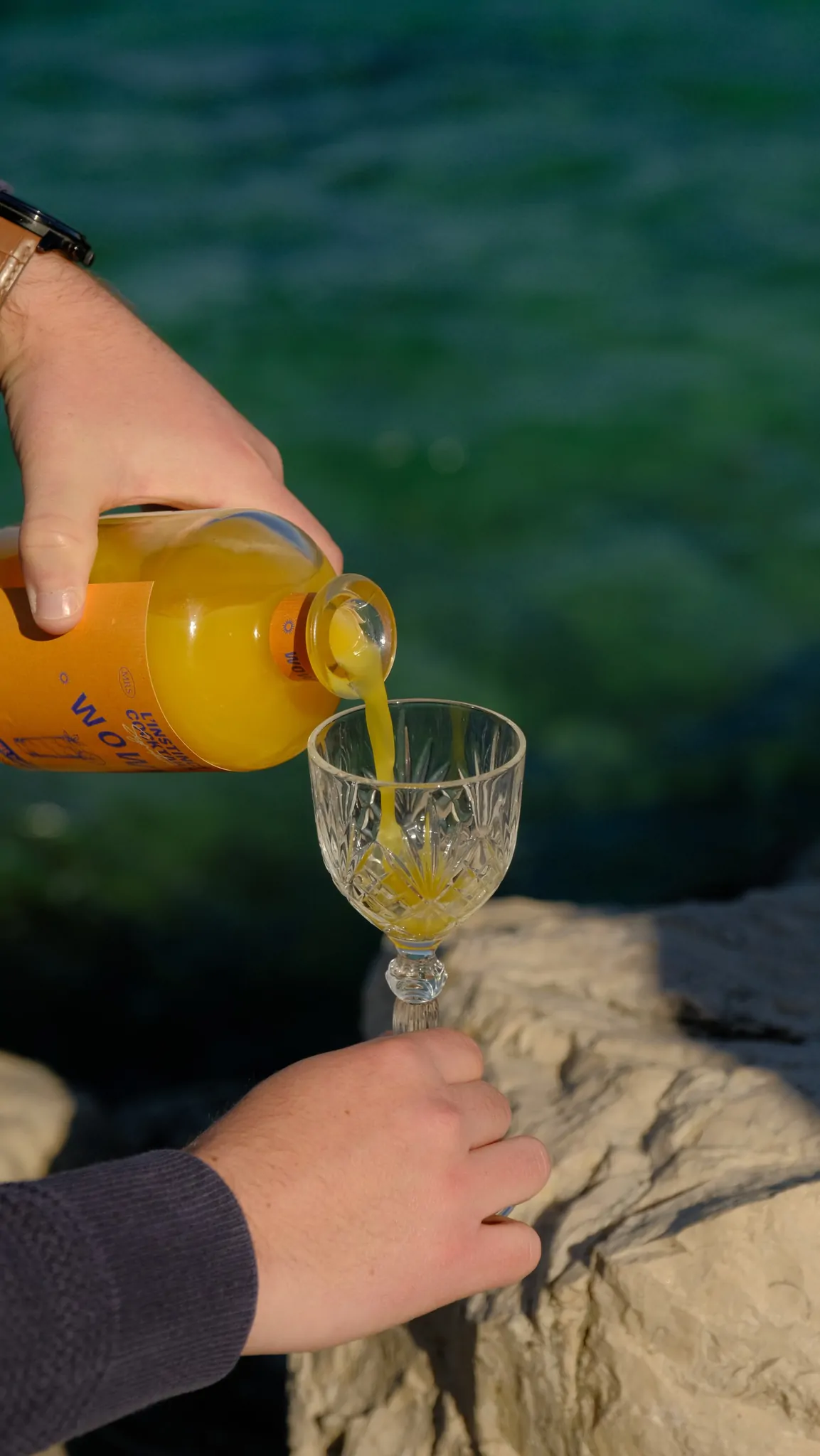 Une main verse le cocktail Wowy d’une bouteille dans un verre en cristal tenu au-dessus d’un rocher, devant l’eau.