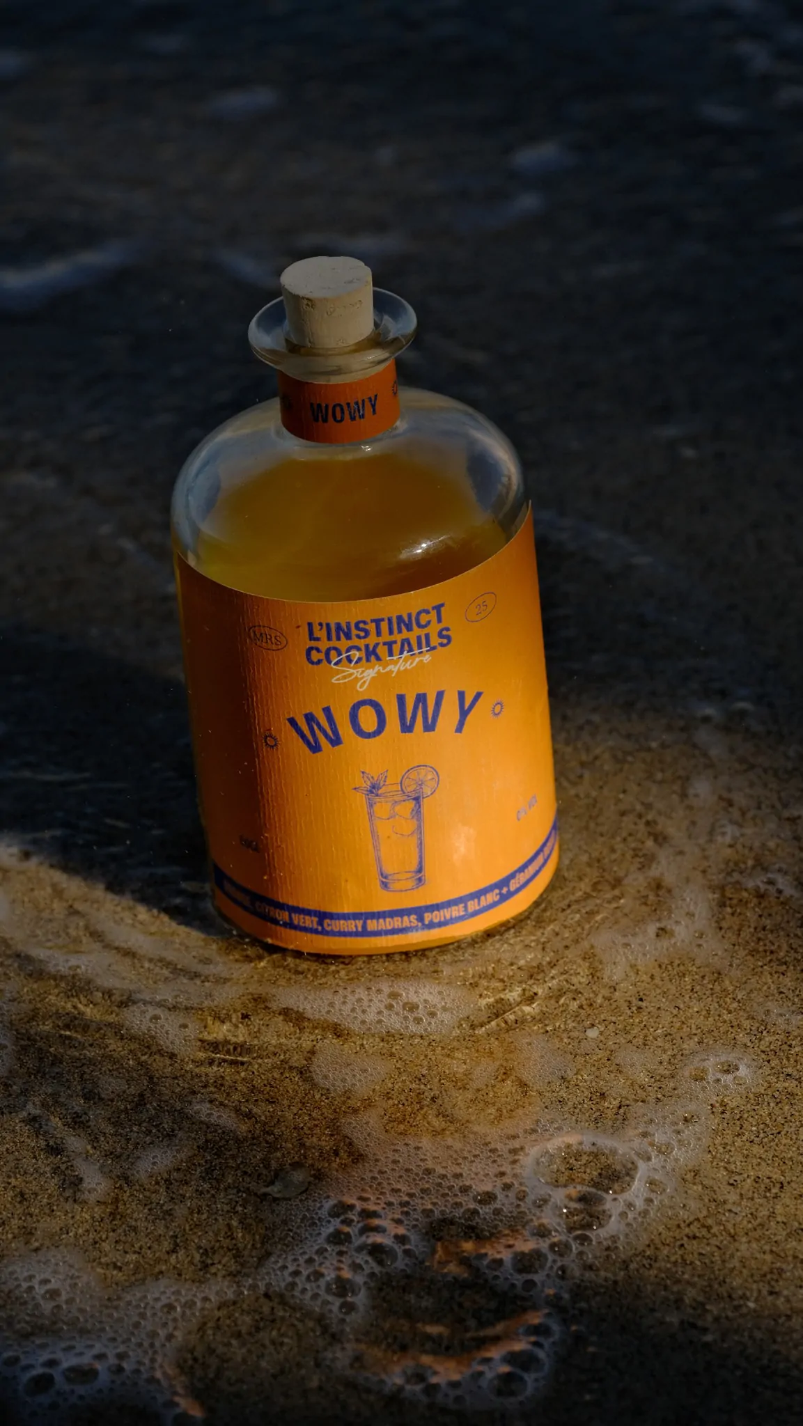 La bouteille Wowy dans l’écume, au bord de l’eau, sur du sable mouillé.