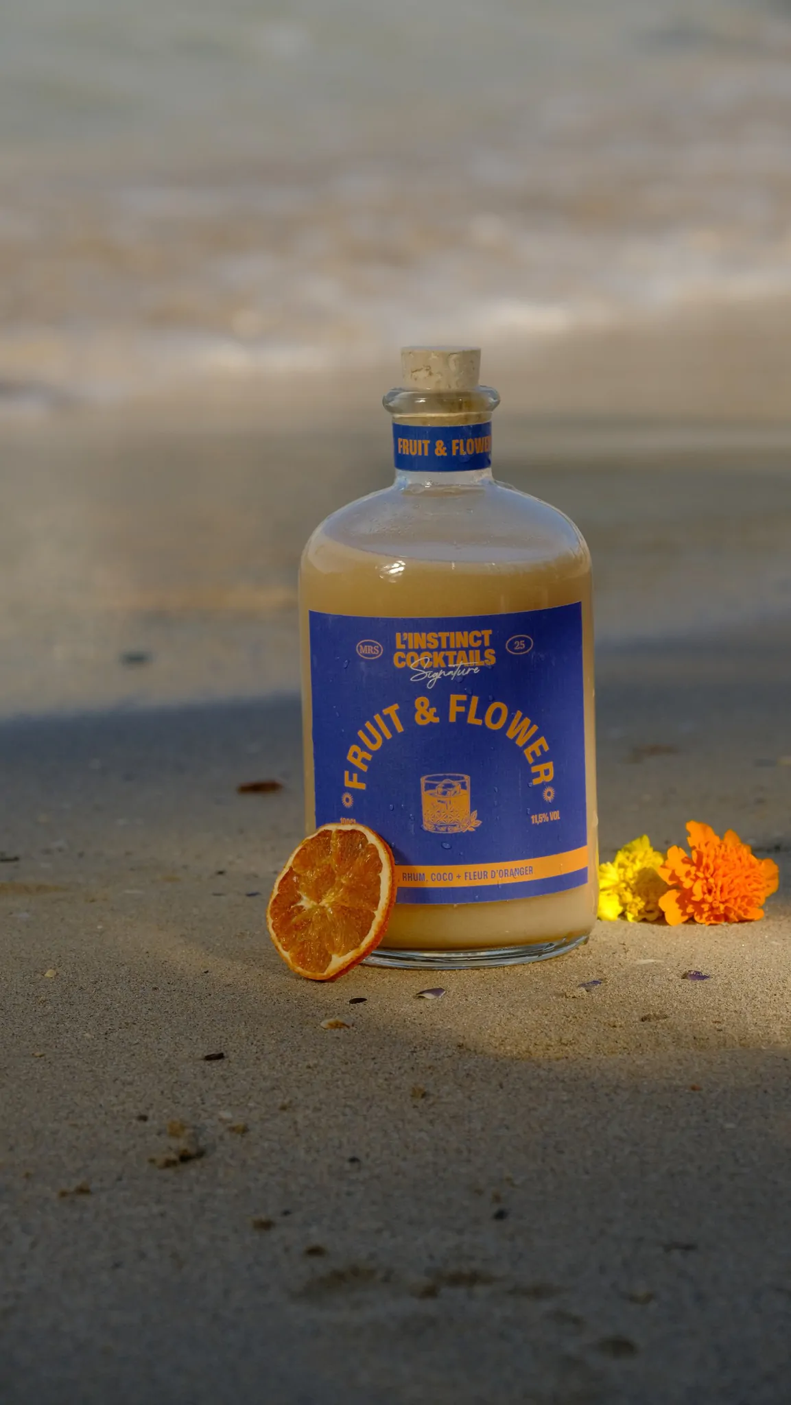 La bouteille Fruit & Flower sur le sable, avec une tranche d’orange séchée et des soucis jaunes, la mer floue derrière.