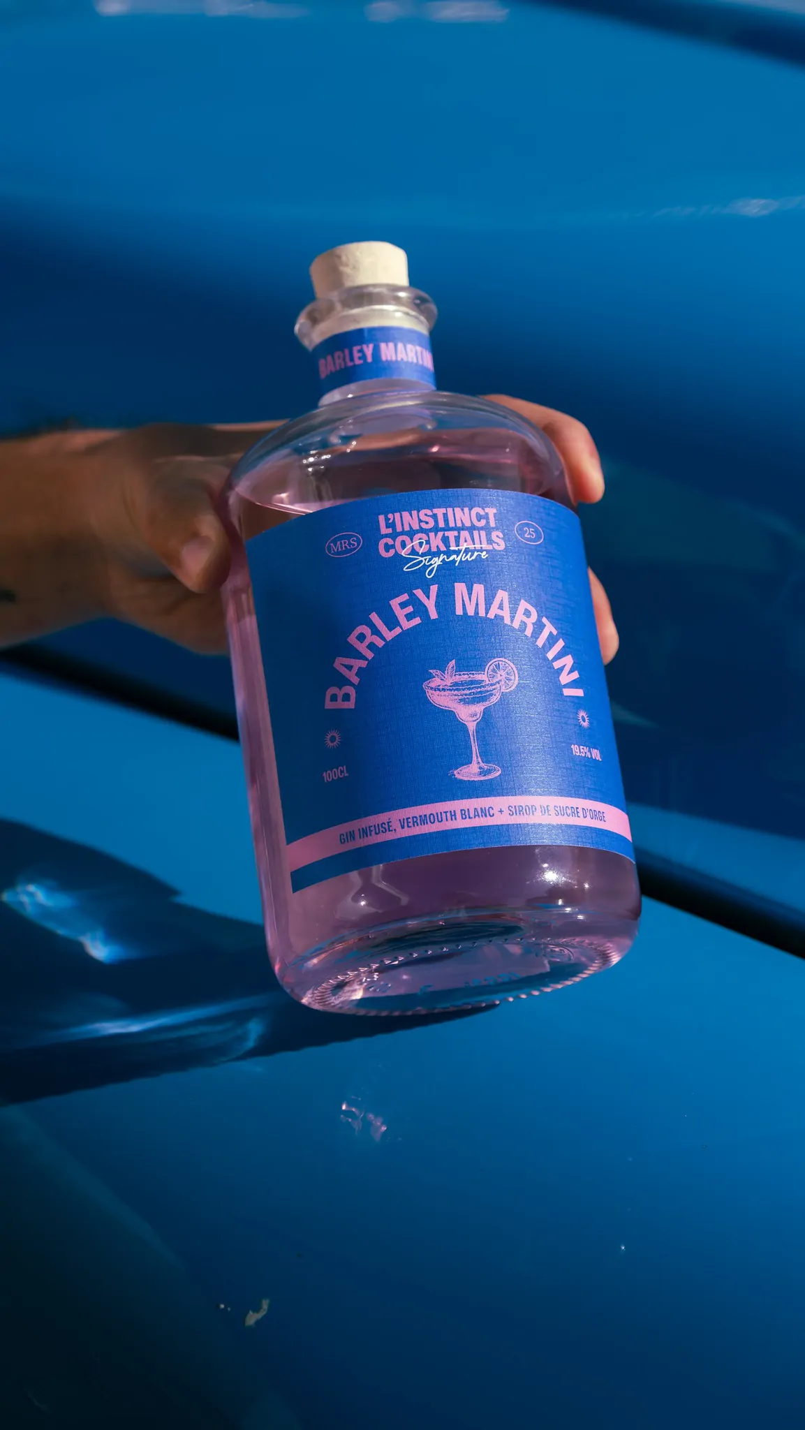 Une main tient la bouteille Barley Martini sur le capot d’une voiture bleue.