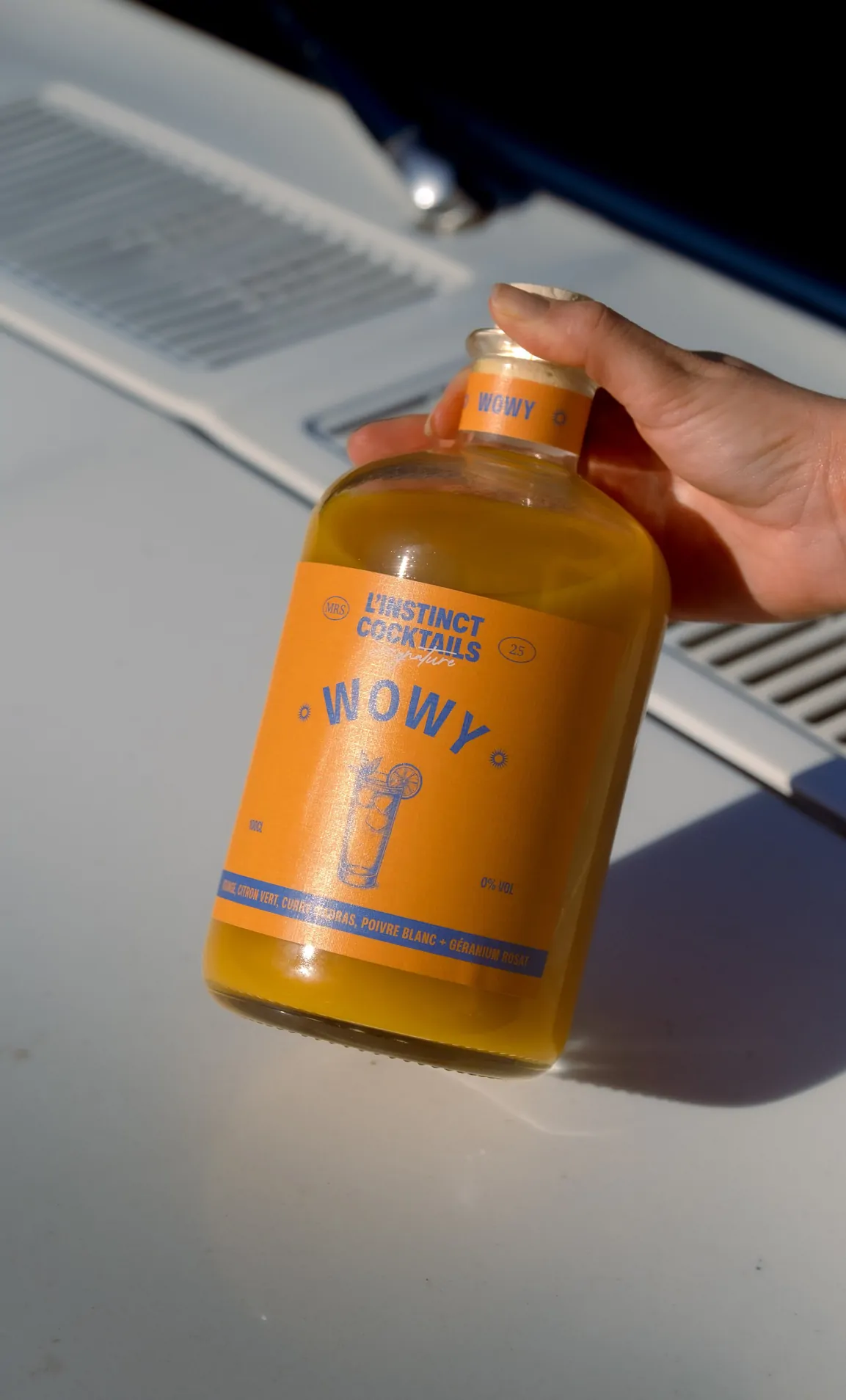 Une main tient la bouteille Wowy sur le capot blanc d’une voiture, près d’une grille d’aération.
