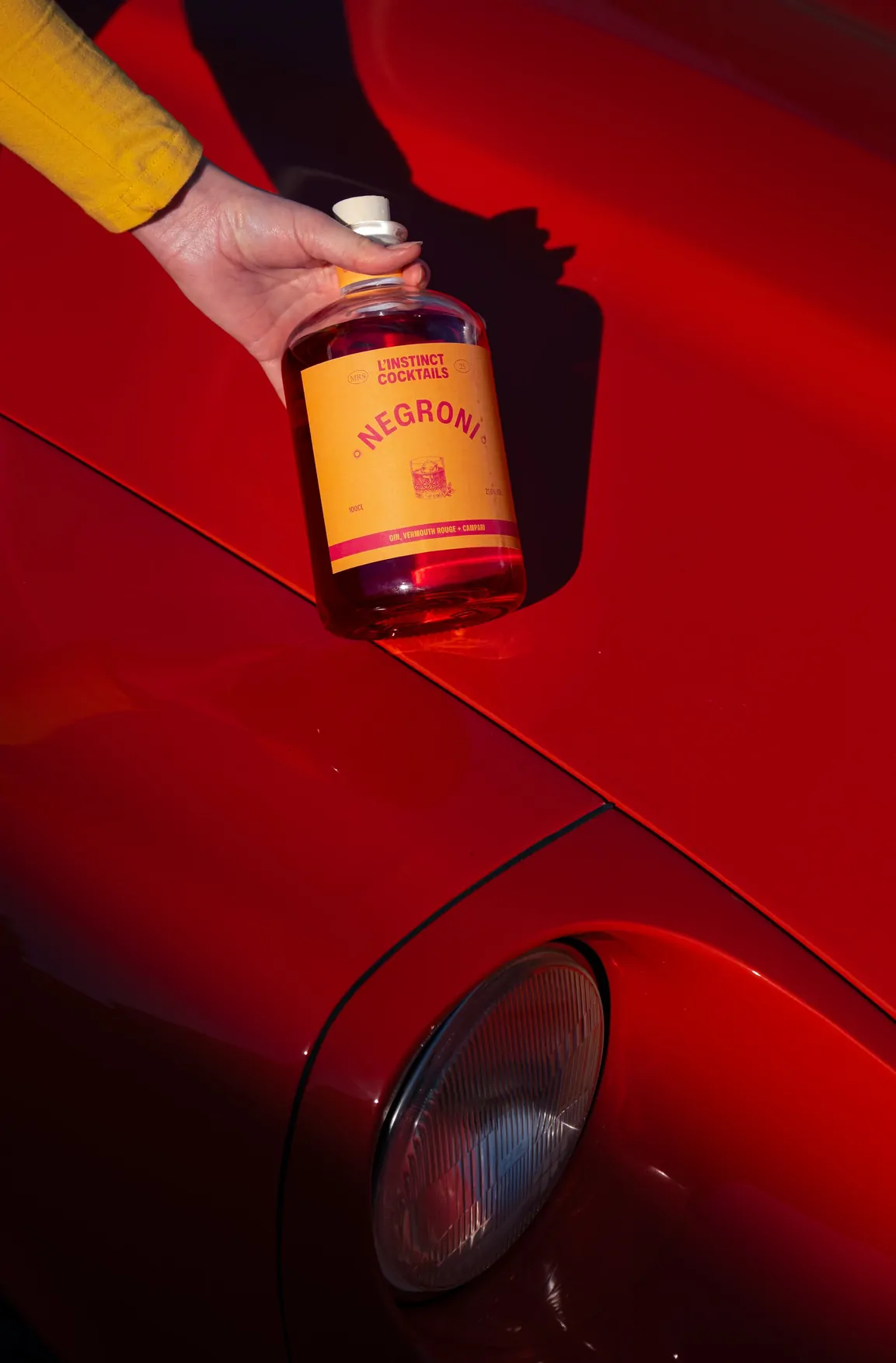 Une main à la manche jaune tient la bouteille Négroni sur l’aile d’une voiture rouge.