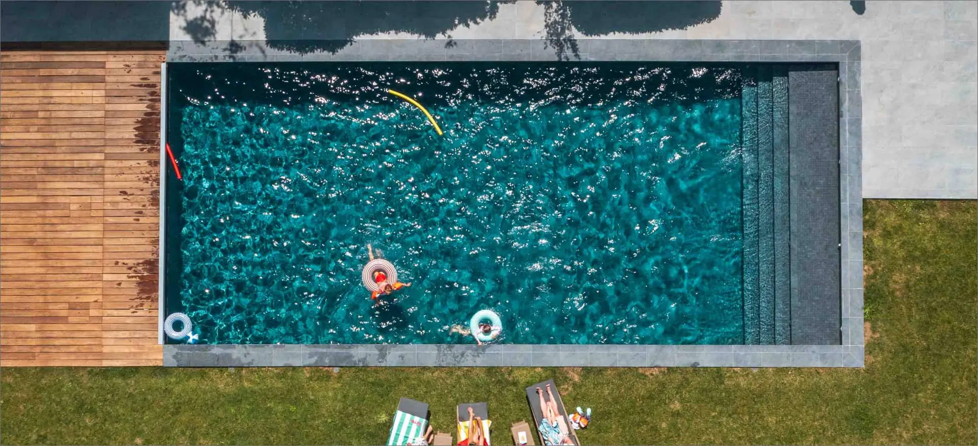 Une piscine rectangulaire vue du ciel, bordée d’une terrasse en bois et d’une pelouse ; une personne sur une bouée, des transats au bord.