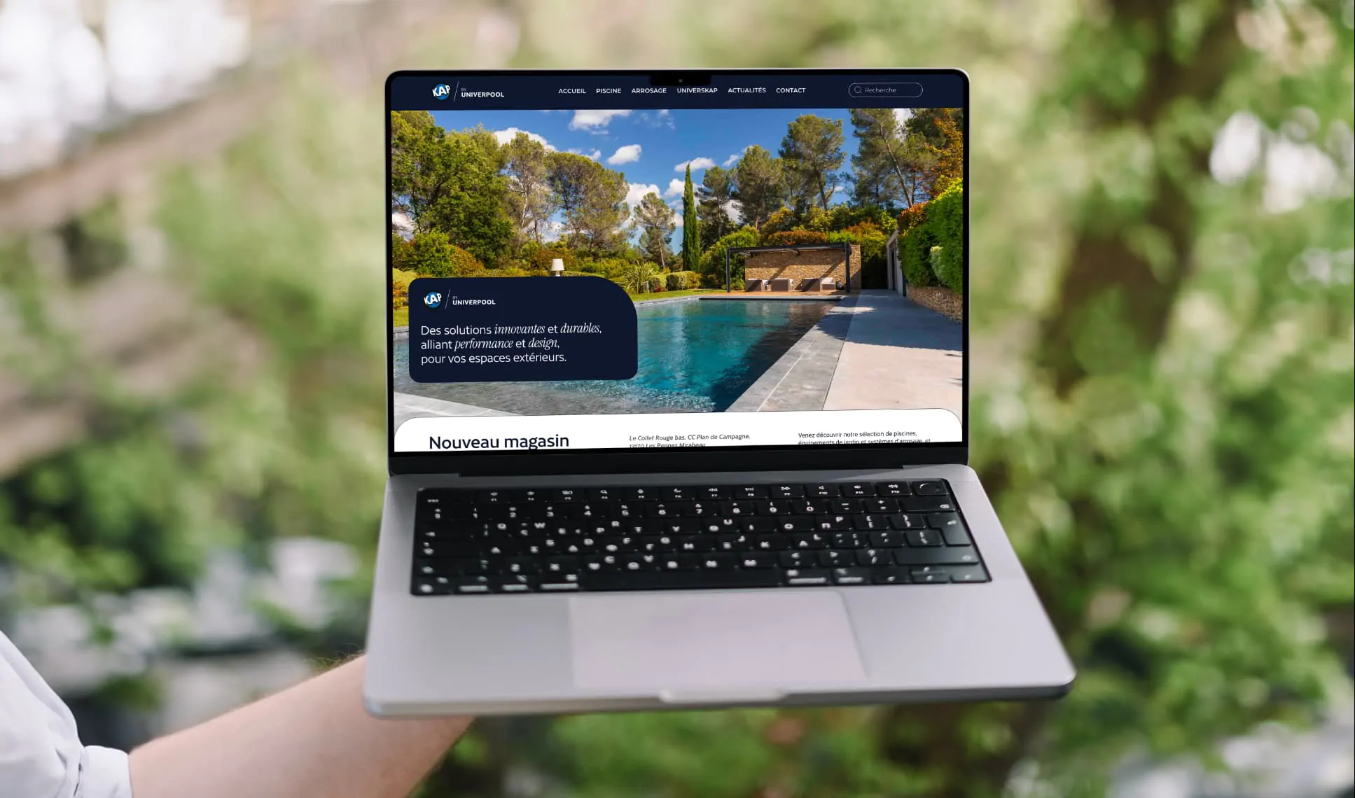 Un ordinateur portable tenu à la main devant un jardin, affichant le site de KAP by Univerpool : une piscine et le titre « Nouveau magasin ».