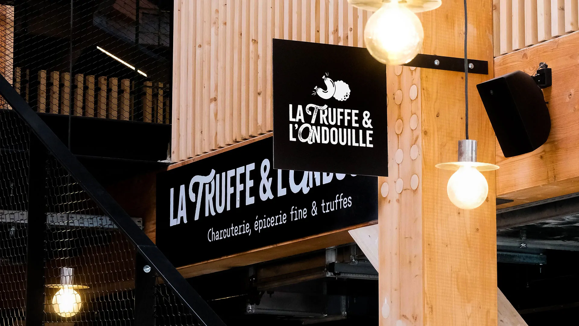 L’enseigne noire « La Truffe & l’Andouille, charcuterie, épicerie fine et truffes », suspendue sous des lampes dans les Halles.