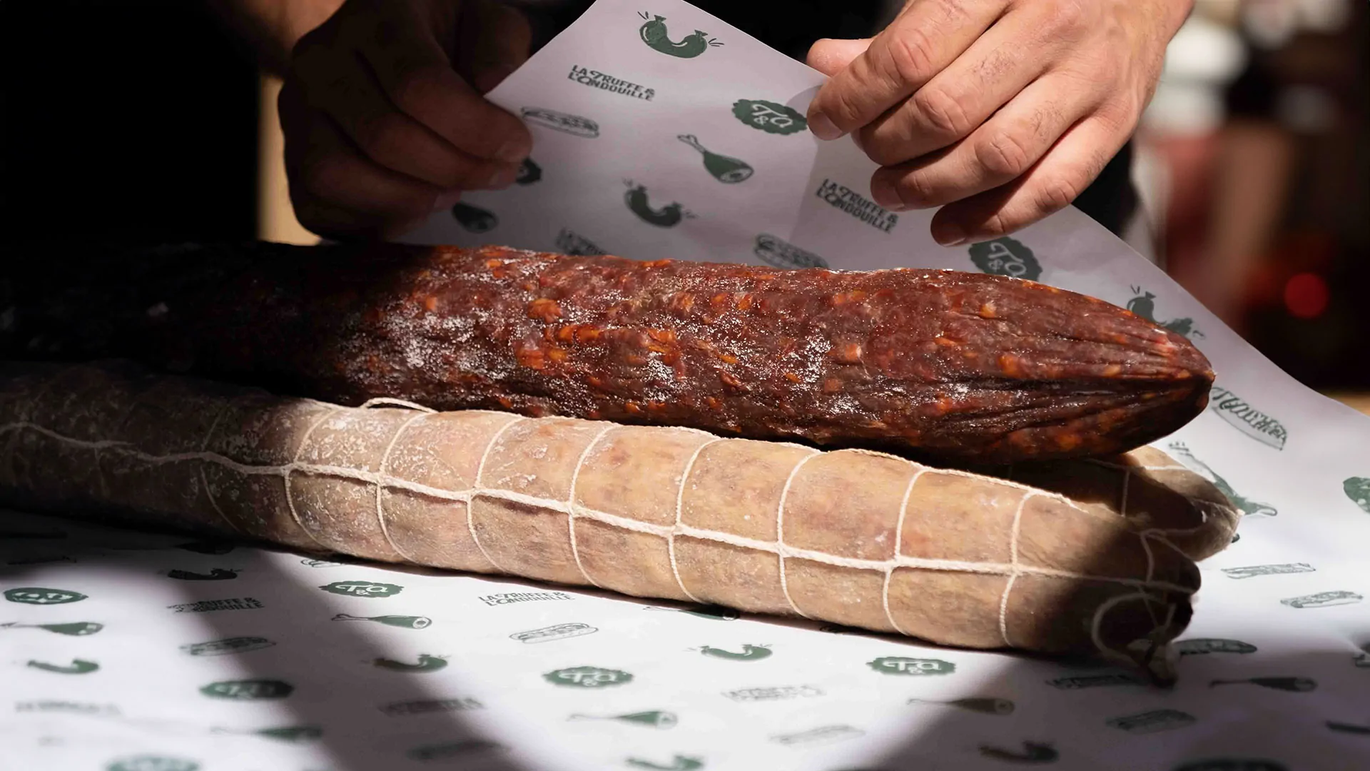Des mains enveloppent un saucisson dans du papier alimentaire à motifs.