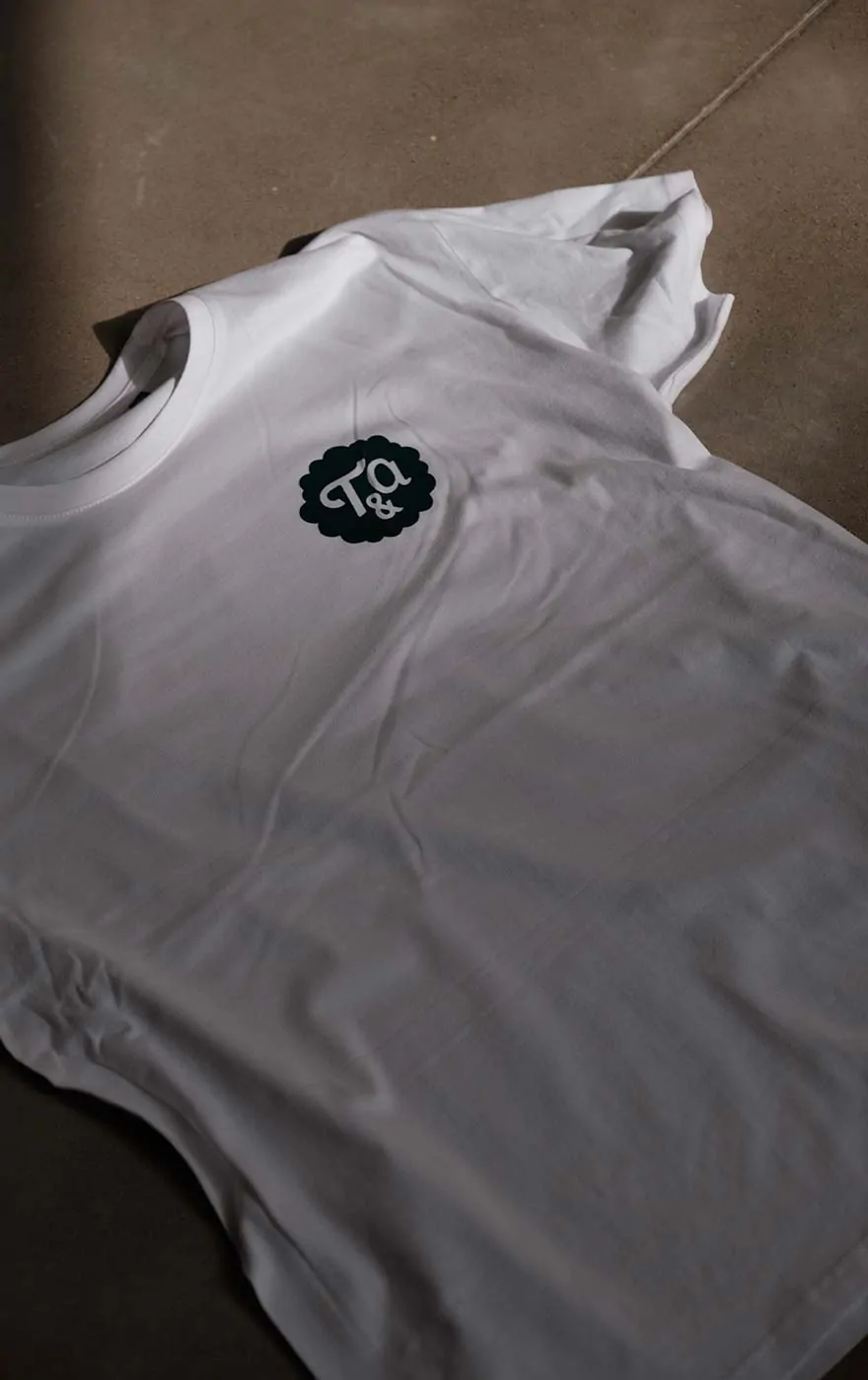 Un tee-shirt blanc orné du badge « T&a » vert sur la poitrine, posé à plat.