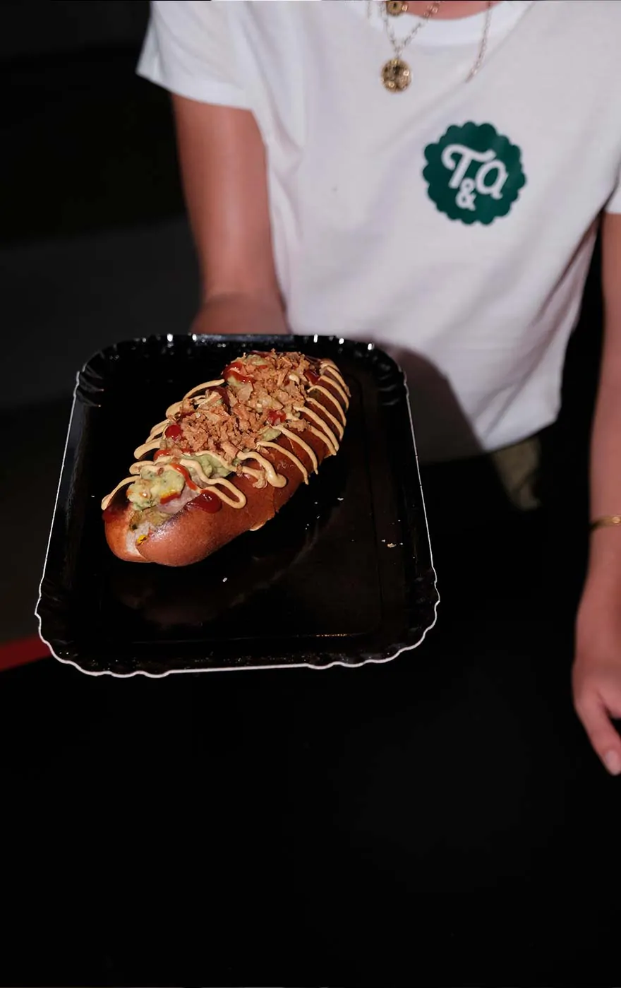 Un plateau noir portant un hot-dog nappé de sauce, tenu devant un tee-shirt blanc au badge vert.