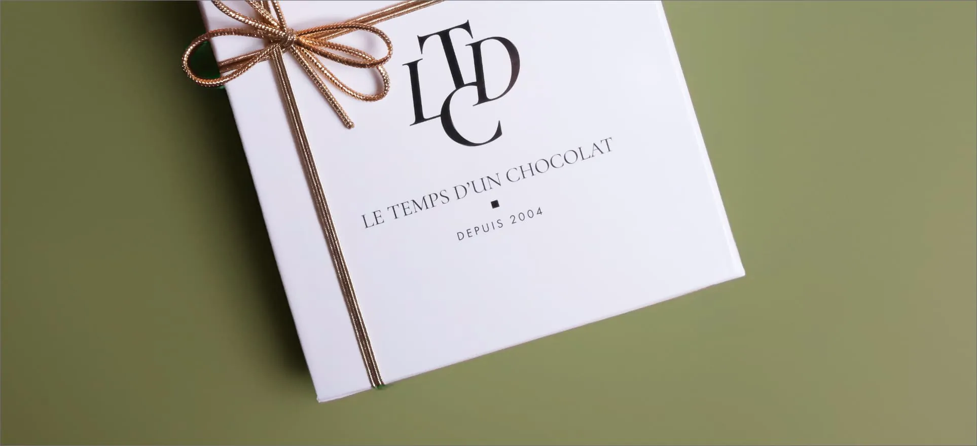 Un coffret blanc noué d’un ruban doré, marqué du monogramme LTDC et de « Le Temps d’un Chocolat, depuis 2004 », posé de biais sur un fond vert olive.