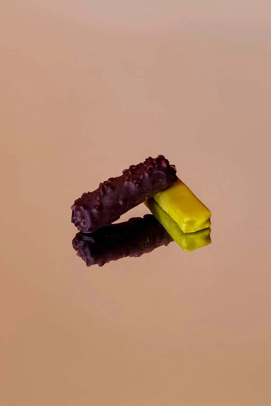 Une barre pistache et une barre au chocolat posées en équilibre sur un fond rose poudré, avec leur reflet.