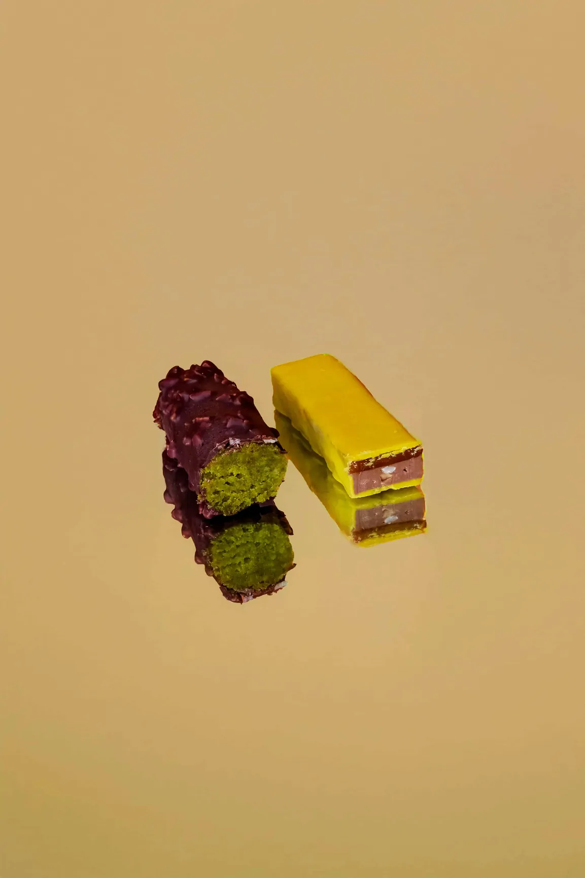 Une barre pistache et une barre au chocolat, dont l’une est coupée en deux, sur un fond jaune sable.
