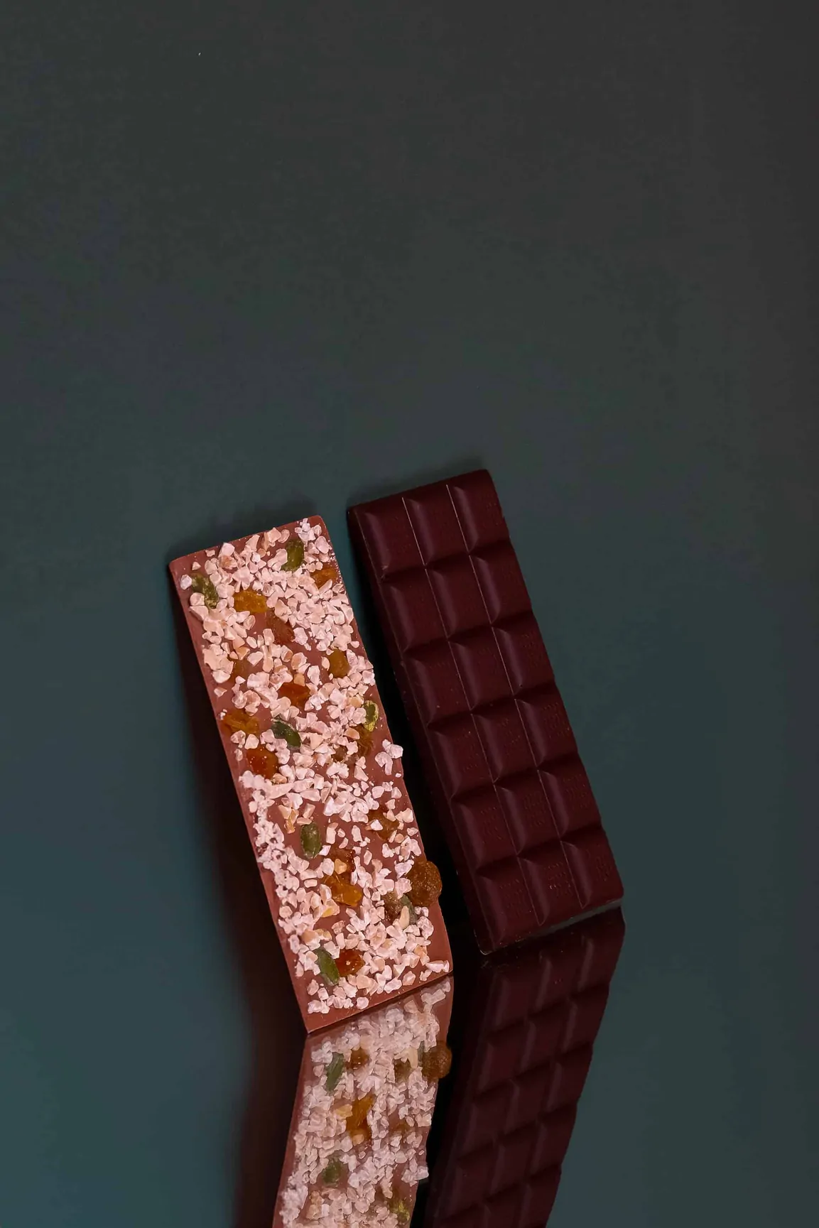 Deux tablettes de chocolat, l’une aux éclats de fruits secs, l’autre noire, sur un fond ardoise, avec leur reflet.