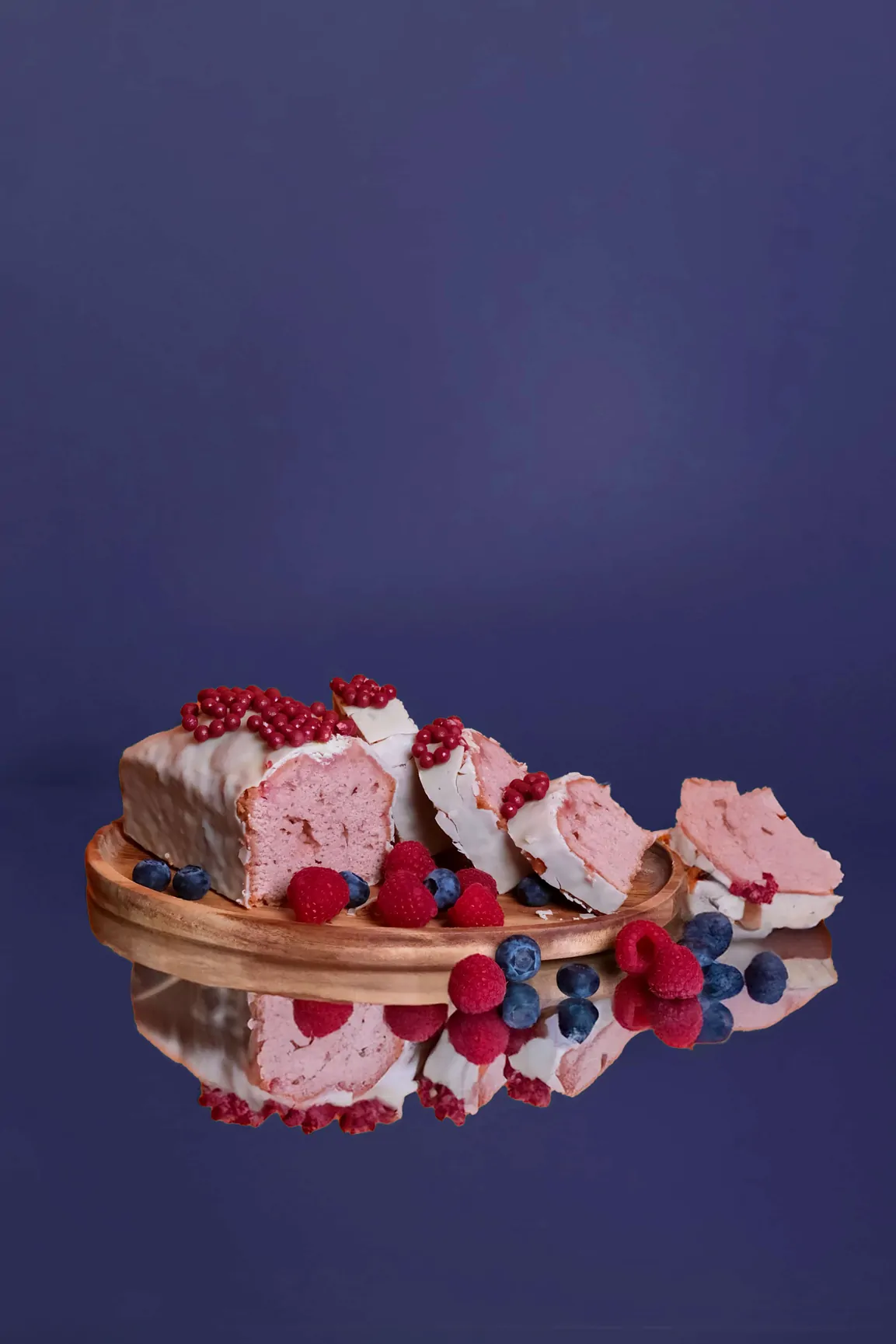 Un gâteau rose aux fruits rouges et aux myrtilles, tranché sur un plateau doré, sur un fond bleu nuit.