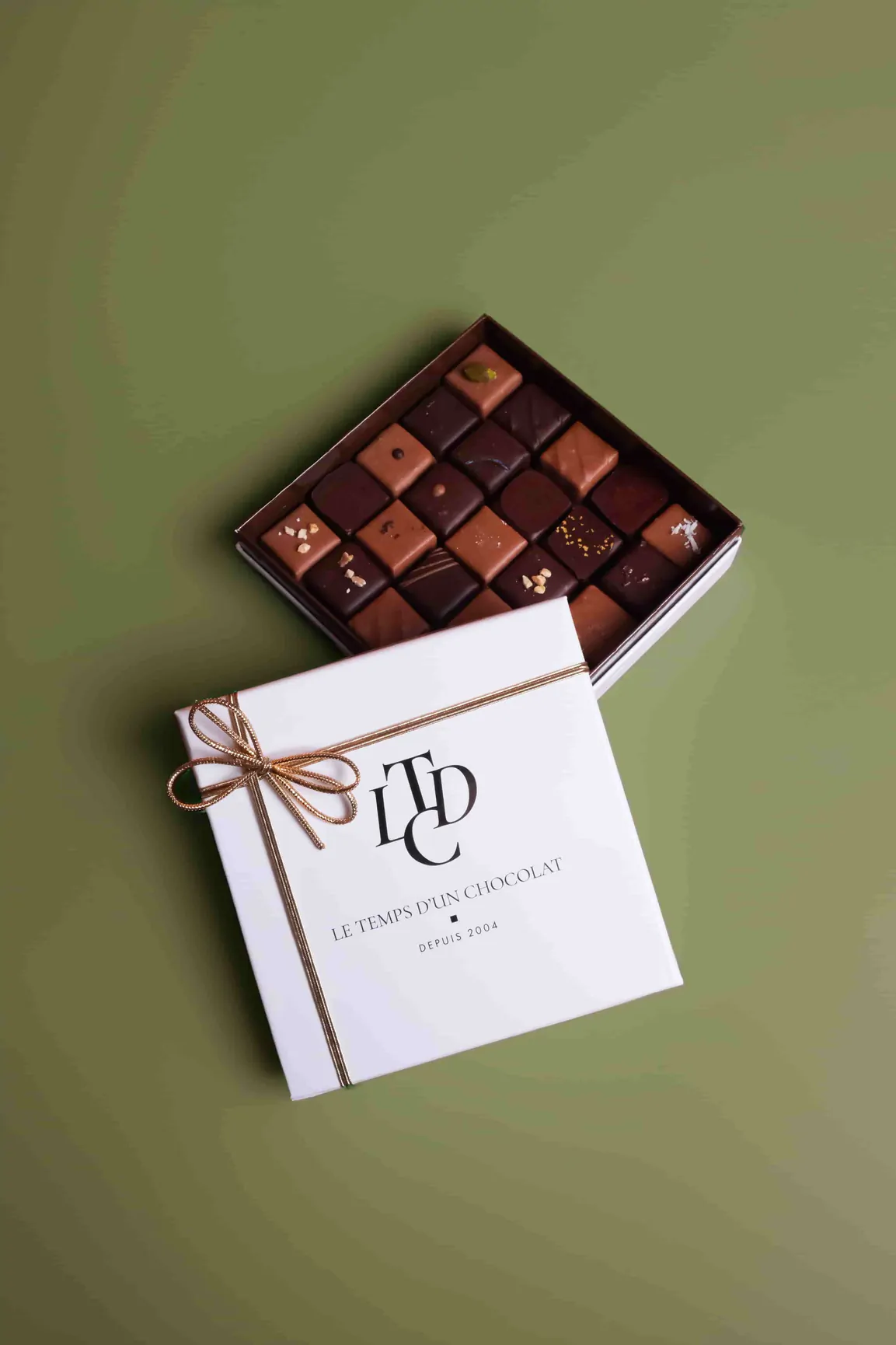 Un coffret ouvert de chocolats carrés garnis, à côté d’un coffret blanc noué d’un ruban doré, sur un fond vert.