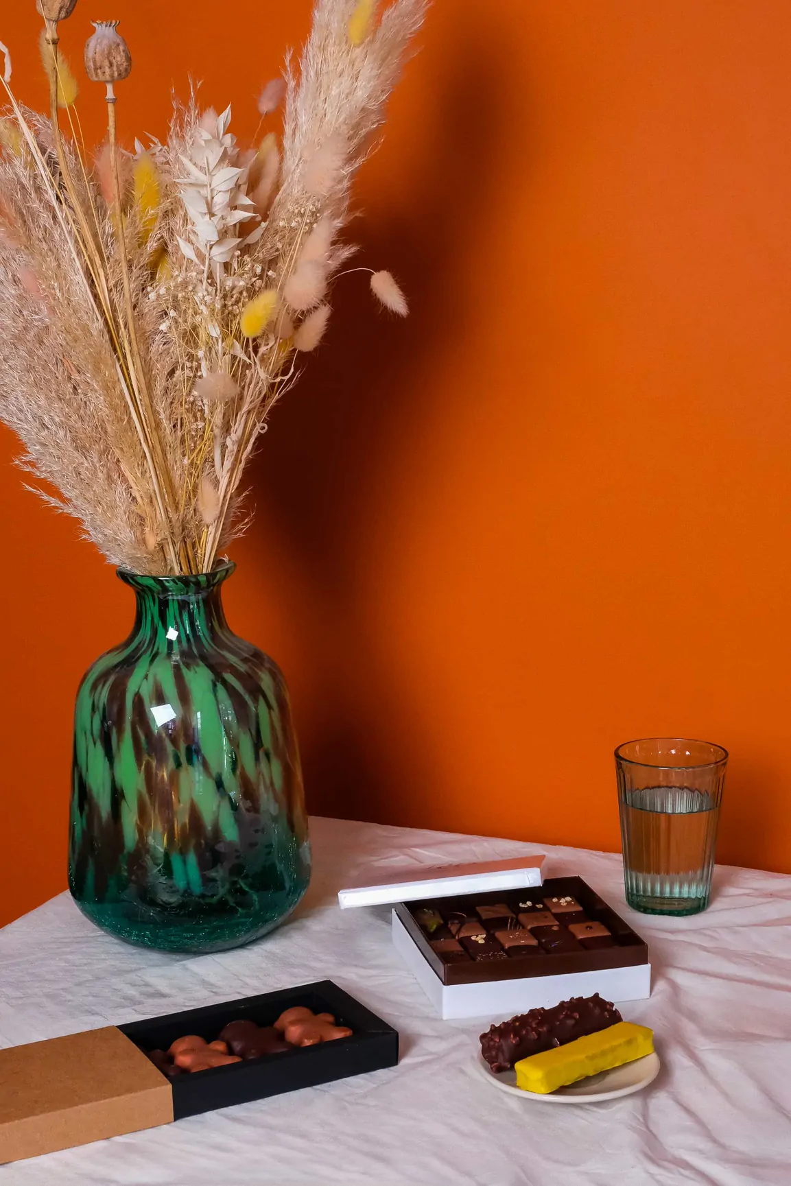 Une table dressée devant un mur orange : un vase vert, un verre d’eau et des boîtes de chocolats.