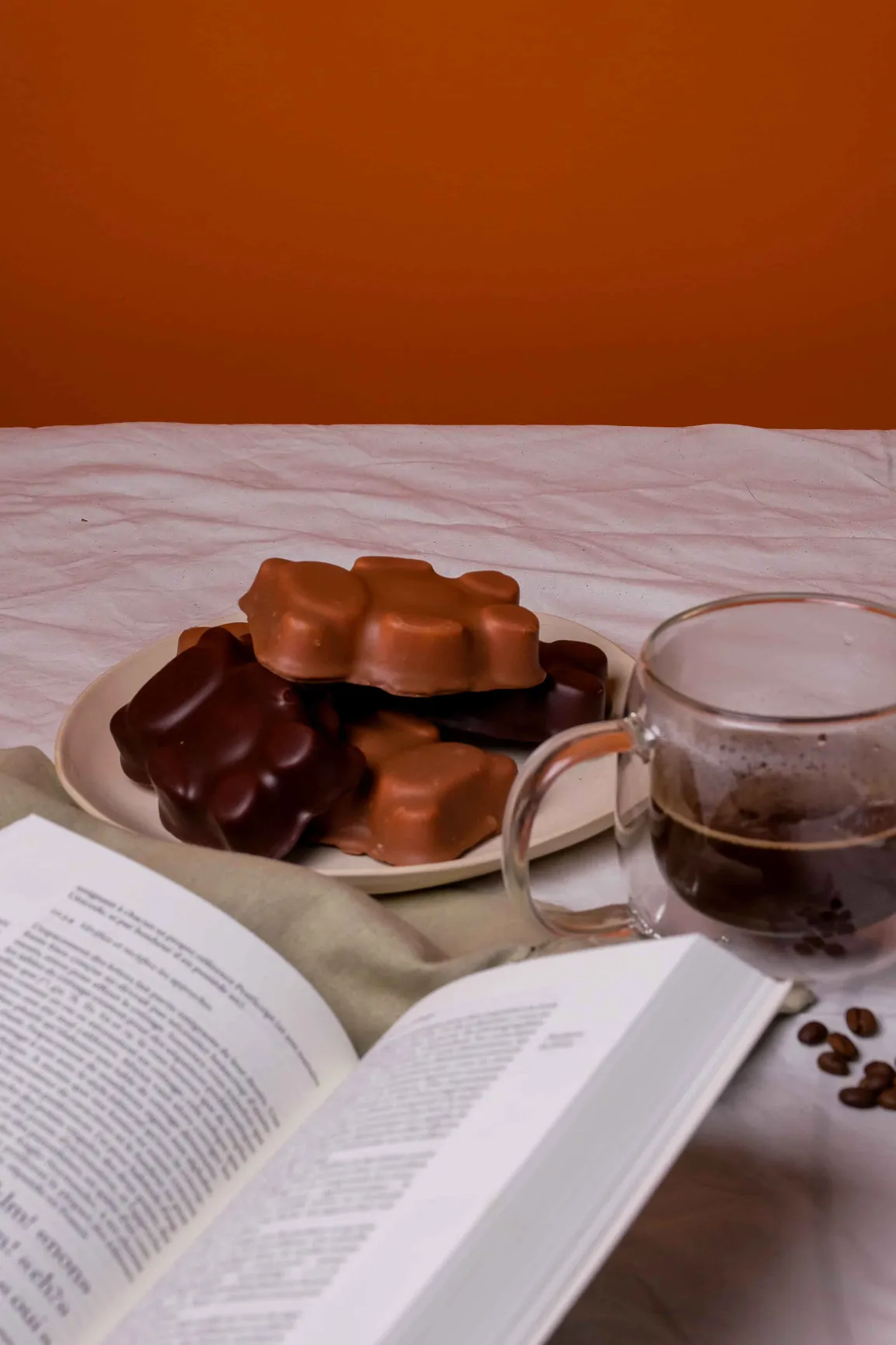 Une assiette de chocolats moulés, une tasse de café et un livre ouvert sur une nappe blanche froissée.