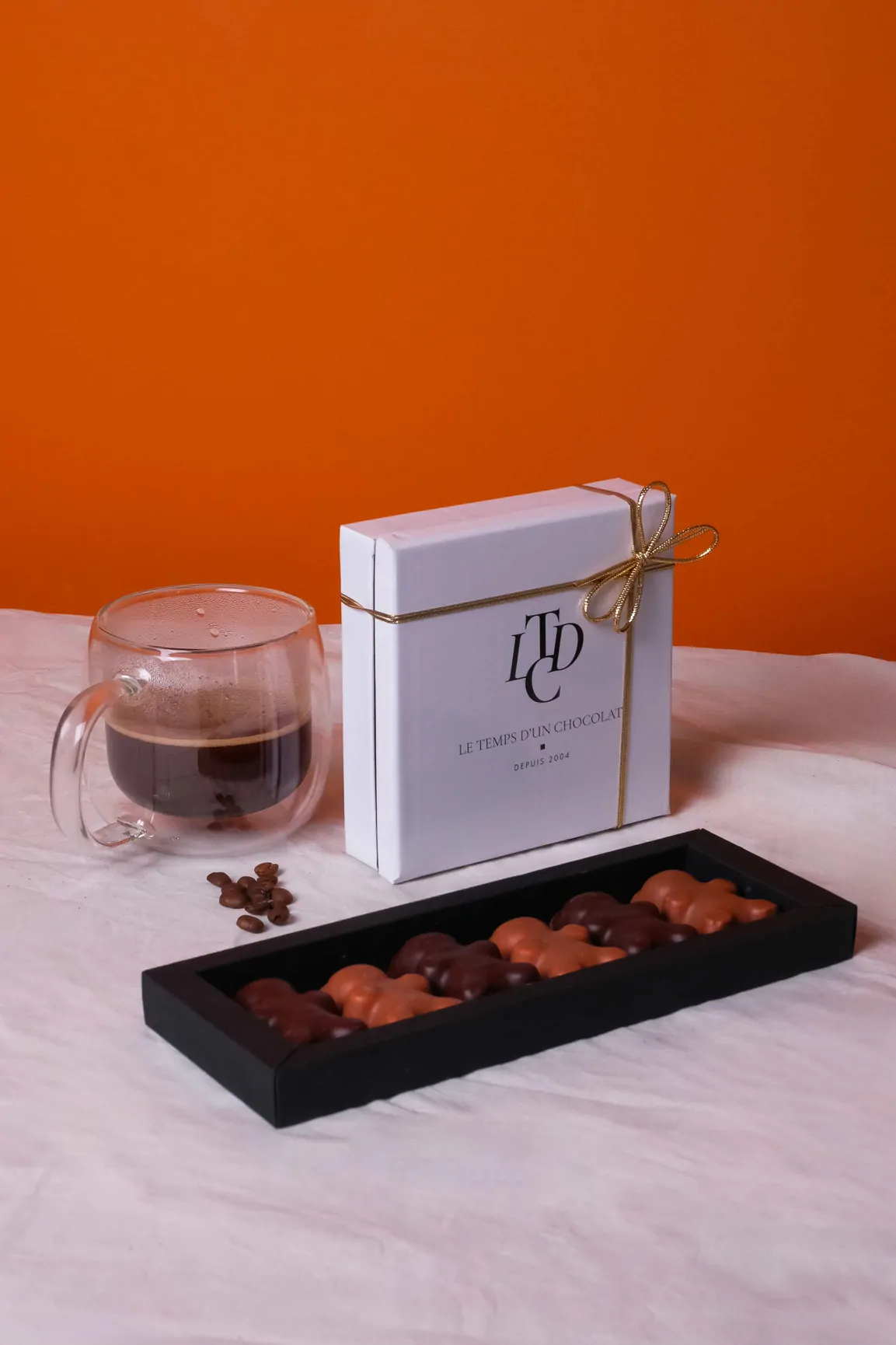 Un coffret blanc au ruban doré, une tasse de café et une boîte noire de chocolats, devant un mur orange.
