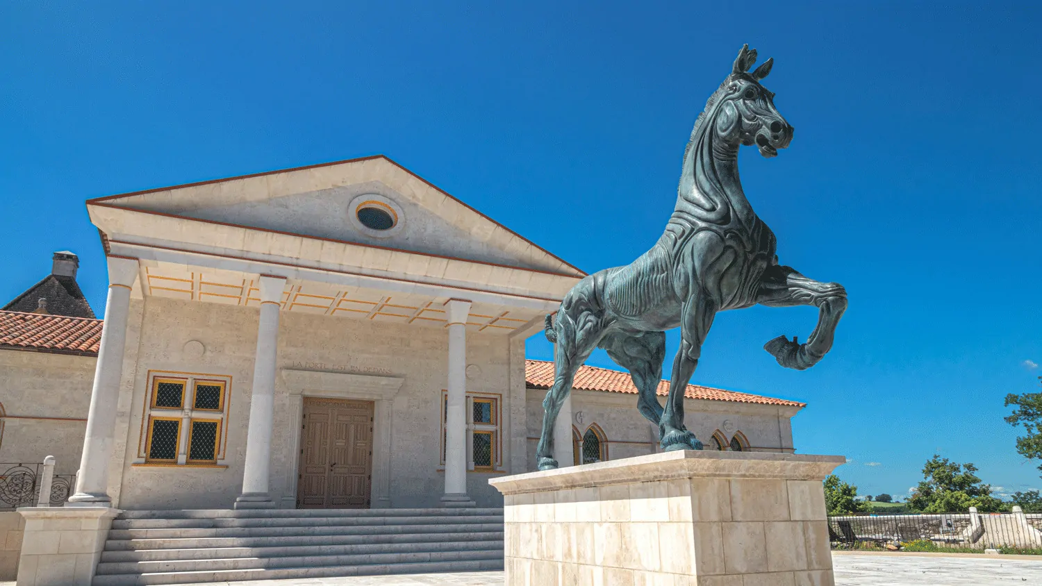 La statue d’un cheval cabré en bronze devant la façade en pierre d’un bâtiment du domaine, sous un ciel bleu.